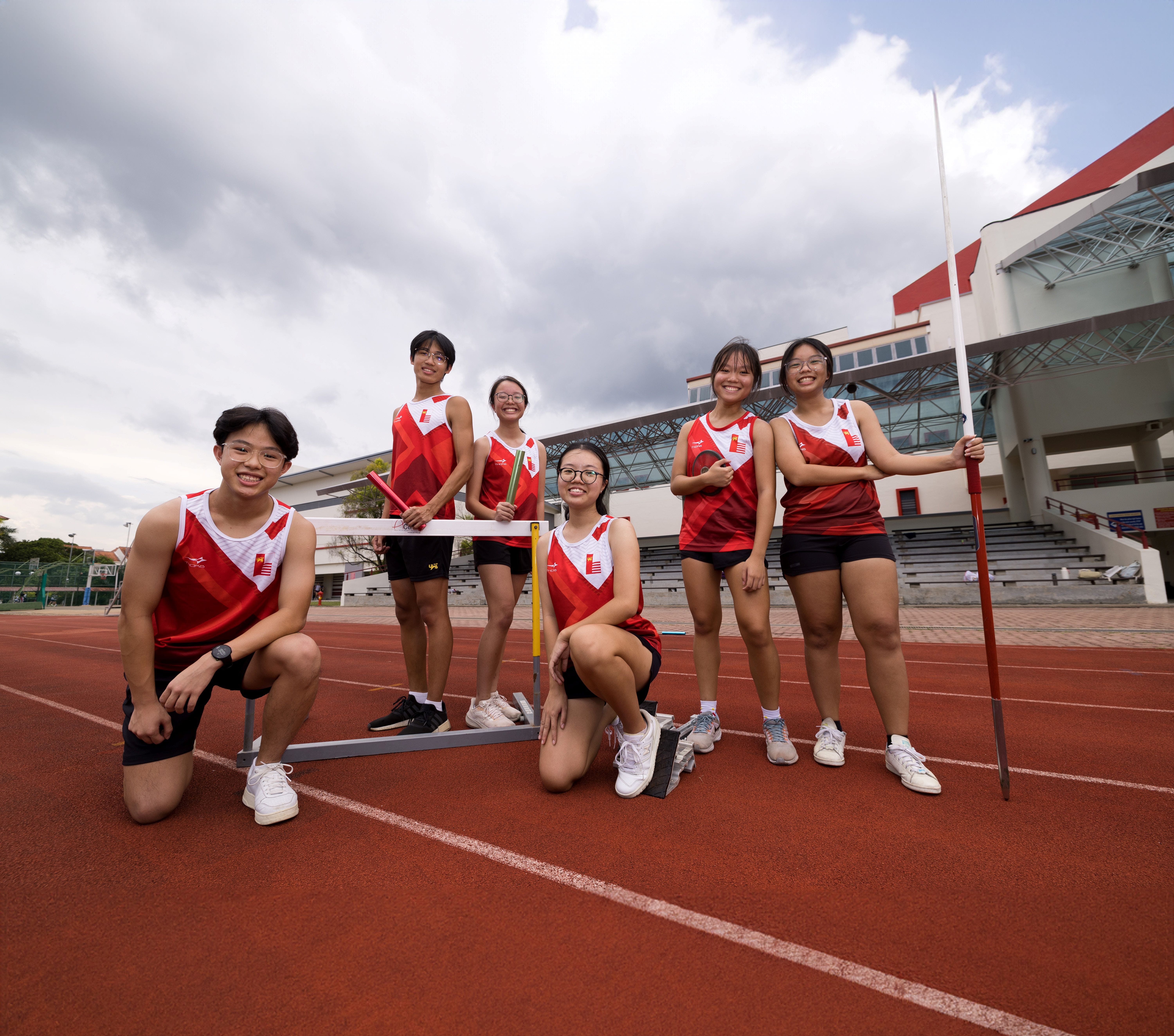 Six young athletes in red uniforms pose on a red running track with a hurdle and javelin.