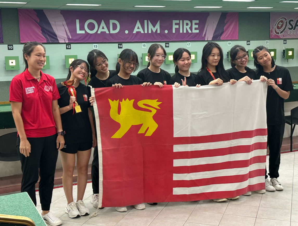 A group of young women hold a red school flag at a shooting range under a "Load. Aim. Fire" banner.