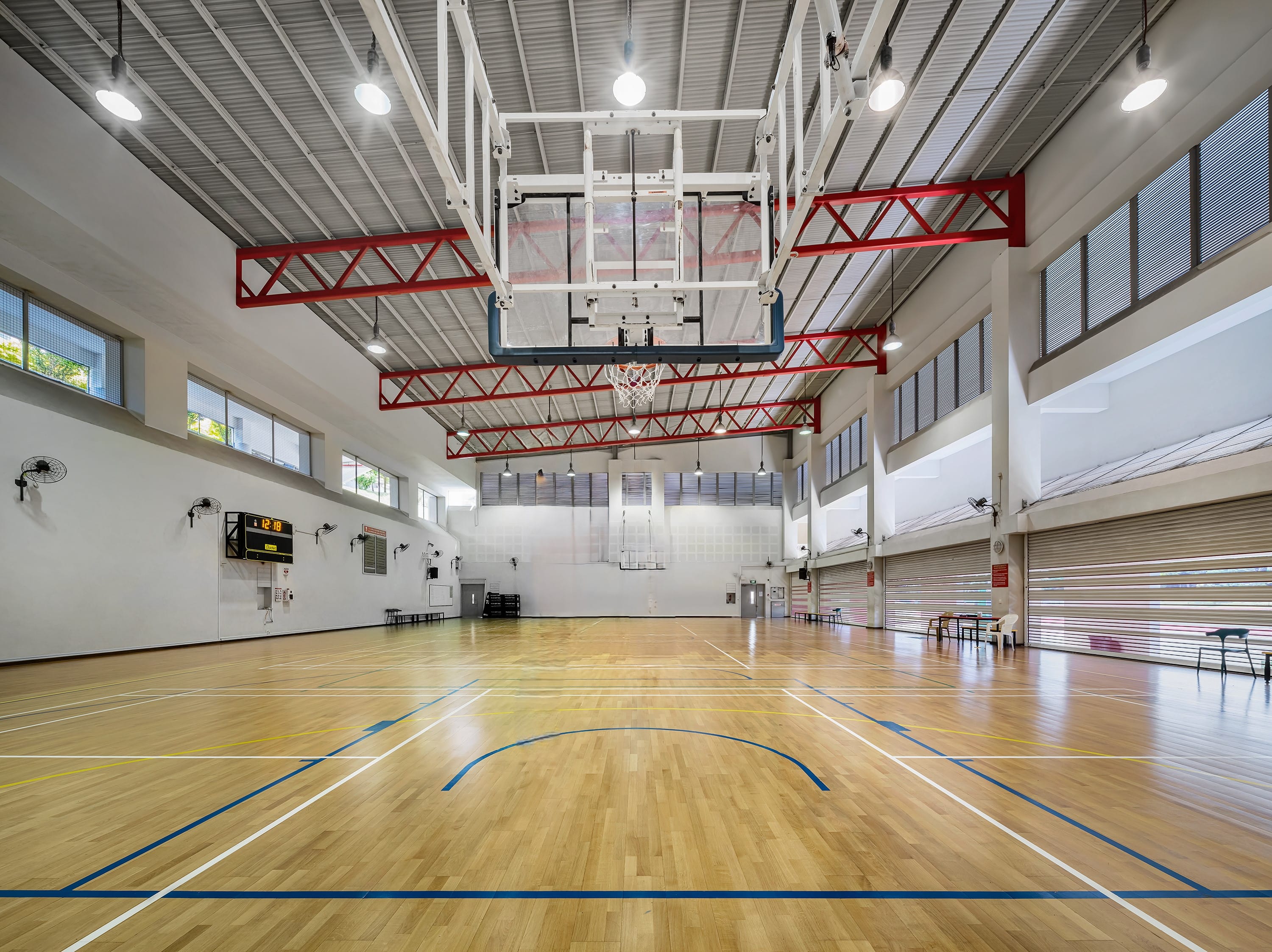 Empty indoor basketball court with a wood floor, overhead hoop, red steel beams, bright lights, and a scoreboard on the wall.