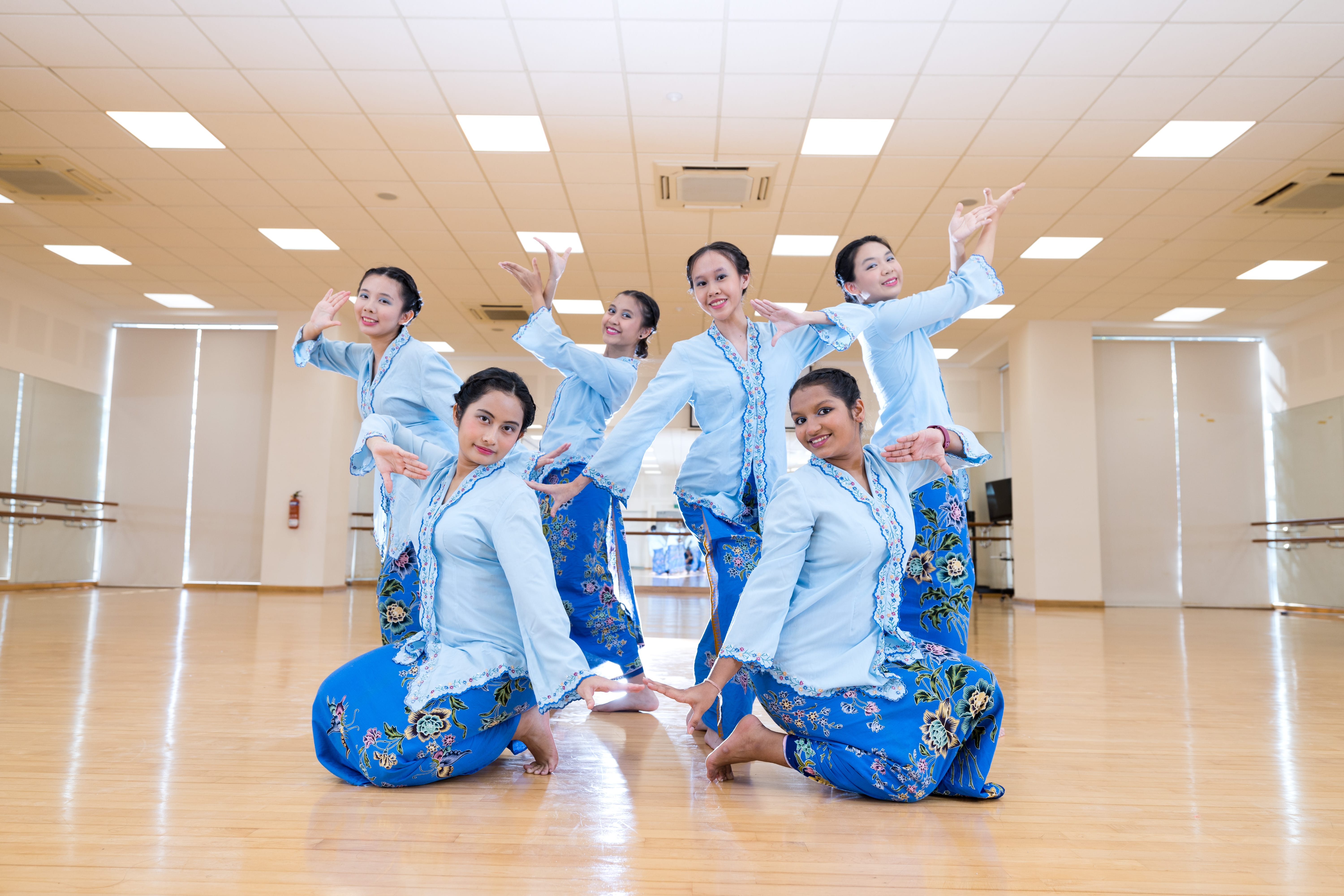 Six dancers in blue outfits pose with raised arms in a bright dance studio with mirrors and wood floor
