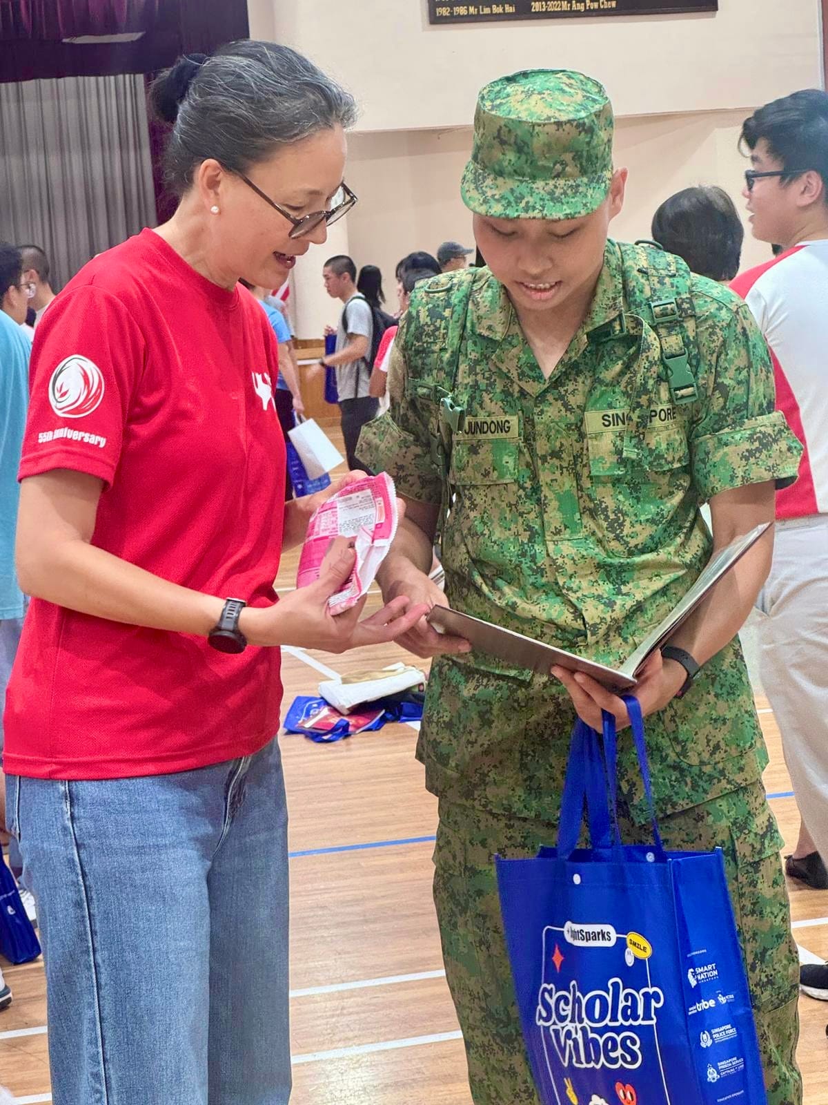 A woman in a red shirt and a Singapore soldier in camouflage look at items together in an indoor hall.