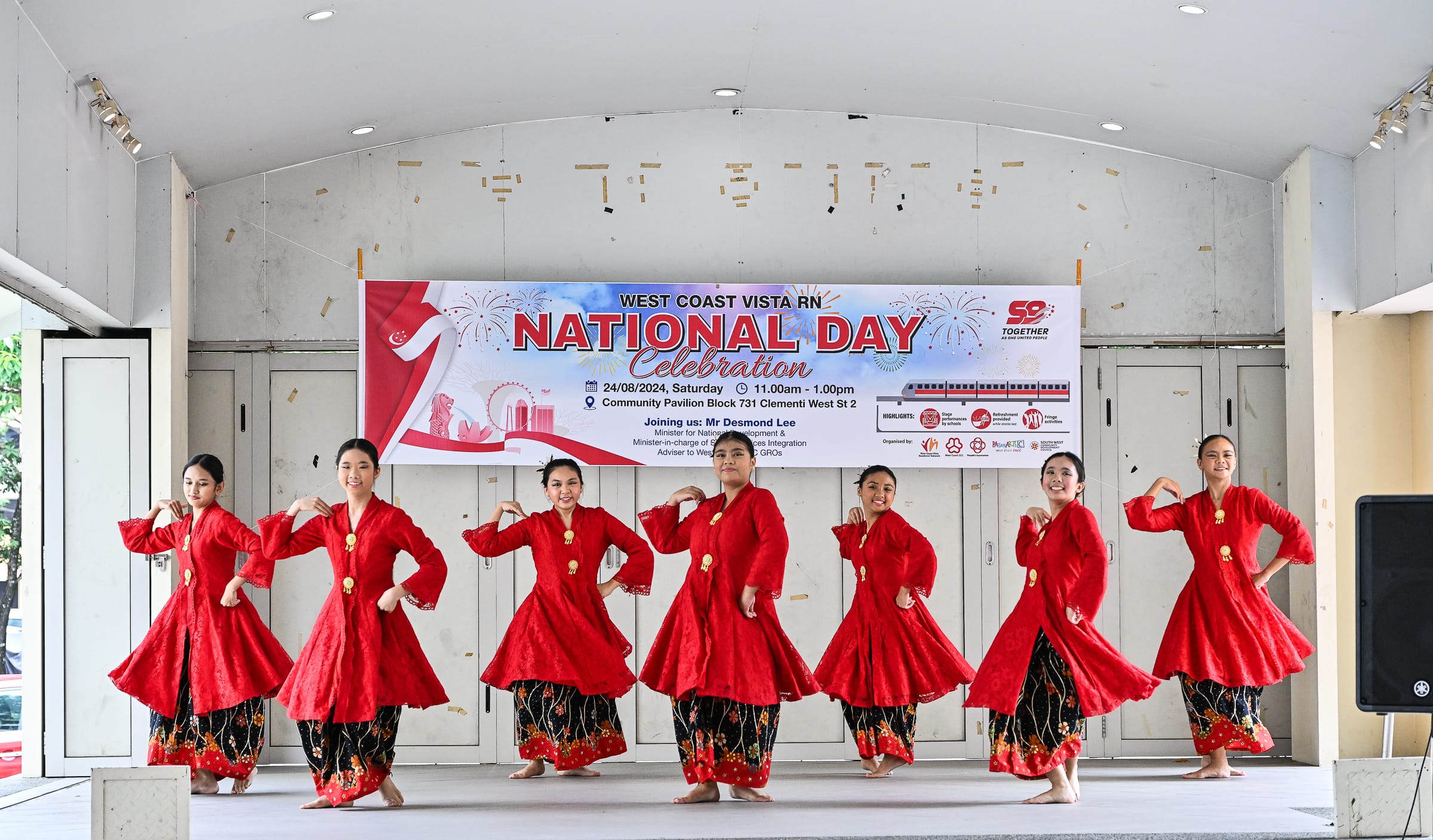 Seven girls in red traditional costumes perform a cultural dance on stage at a National Day Celebration event.
