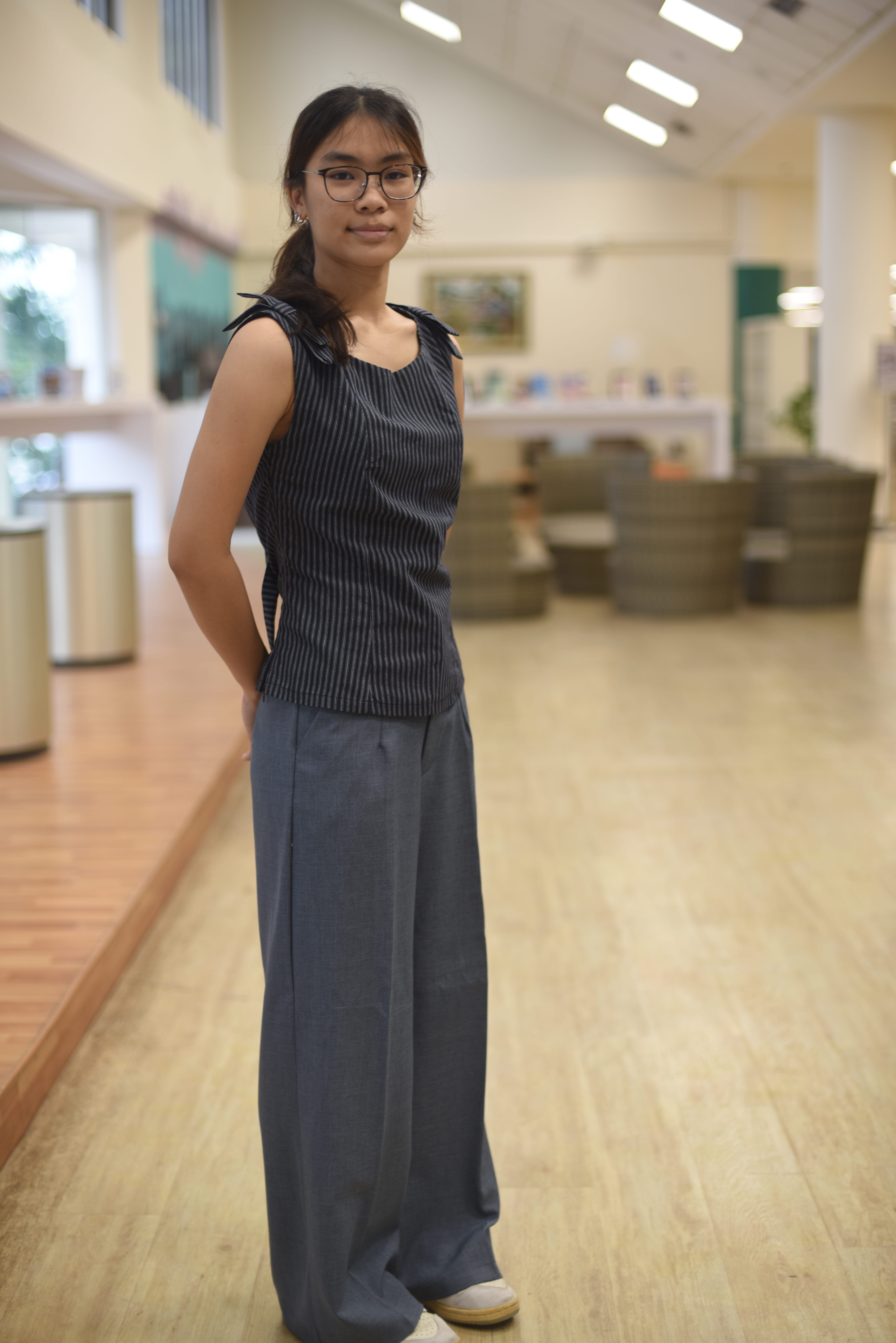 Person with glasses in a sleeveless striped top and wide-leg pants standing in a bright lobby.