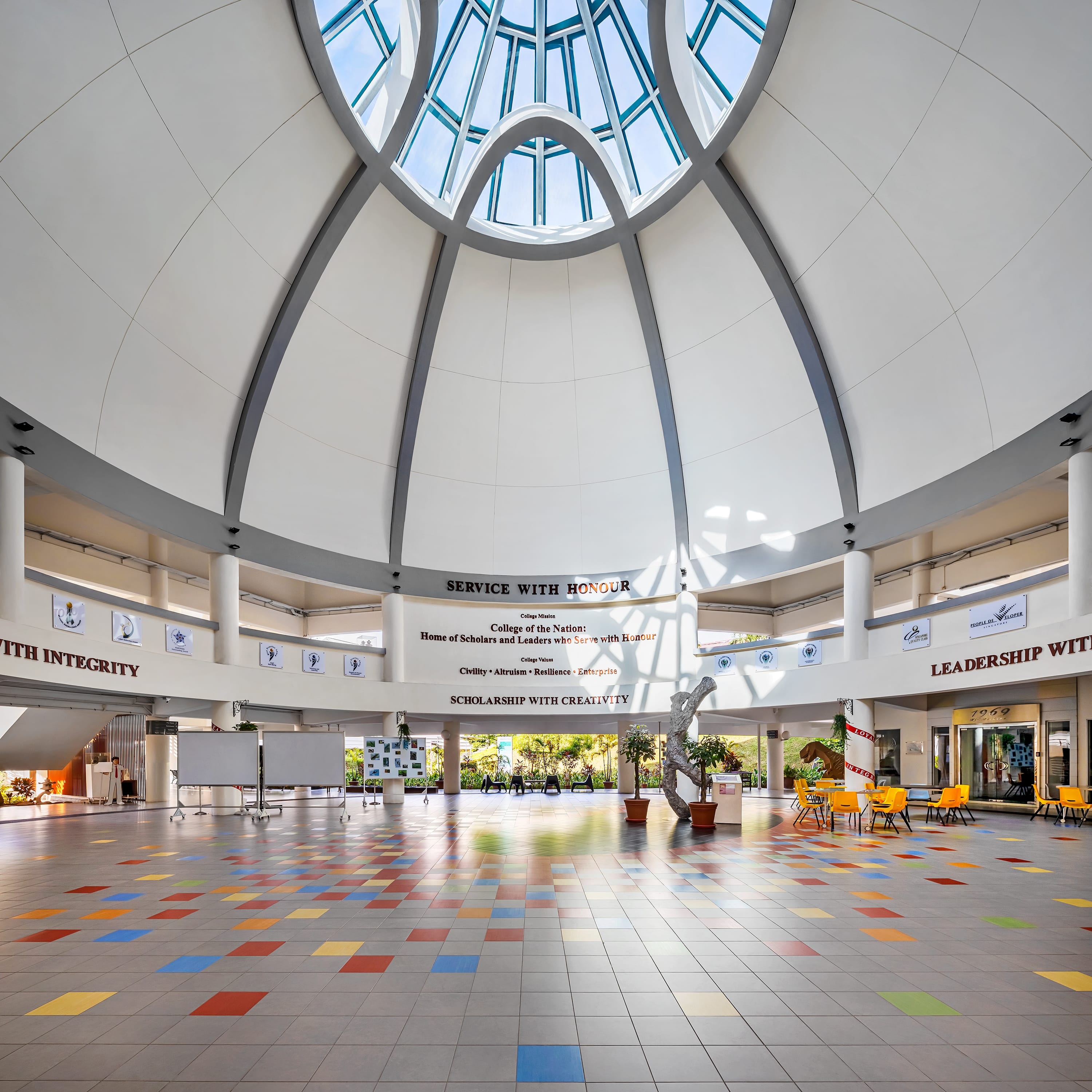 Bright atrium with domed skylight, colorful tiled floor, wall banners, and tables with yellow chairs.