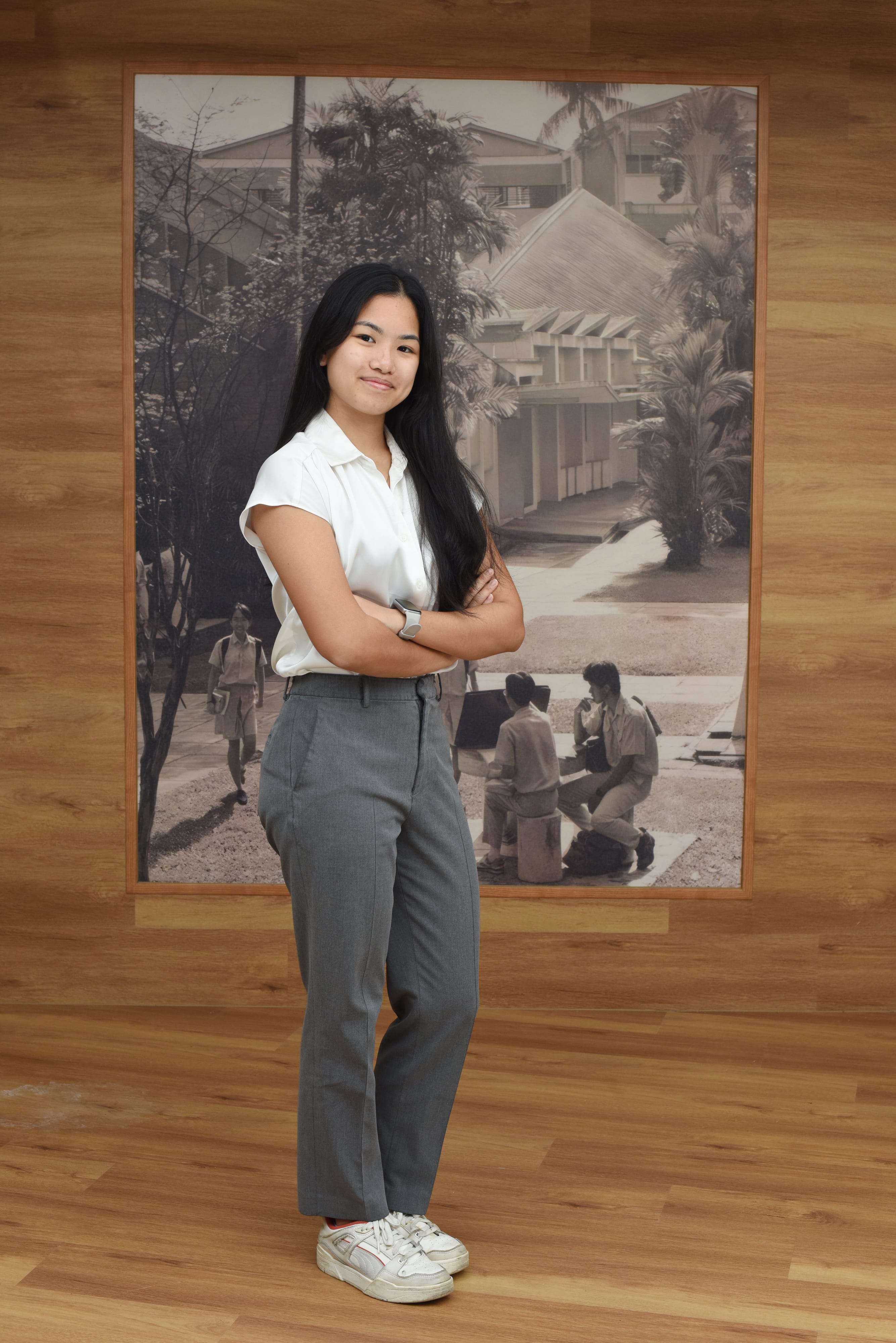 Young woman with long black hair, arms crossed, wearing white shirt and gray pants, standing before a sepia school photo.