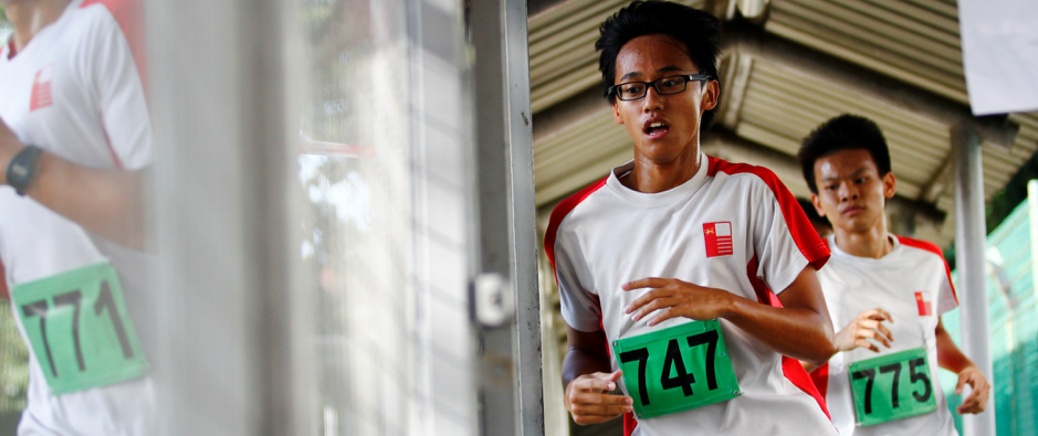Two young male runners wearing green race bibs numbered 747 and 775 run past a building.