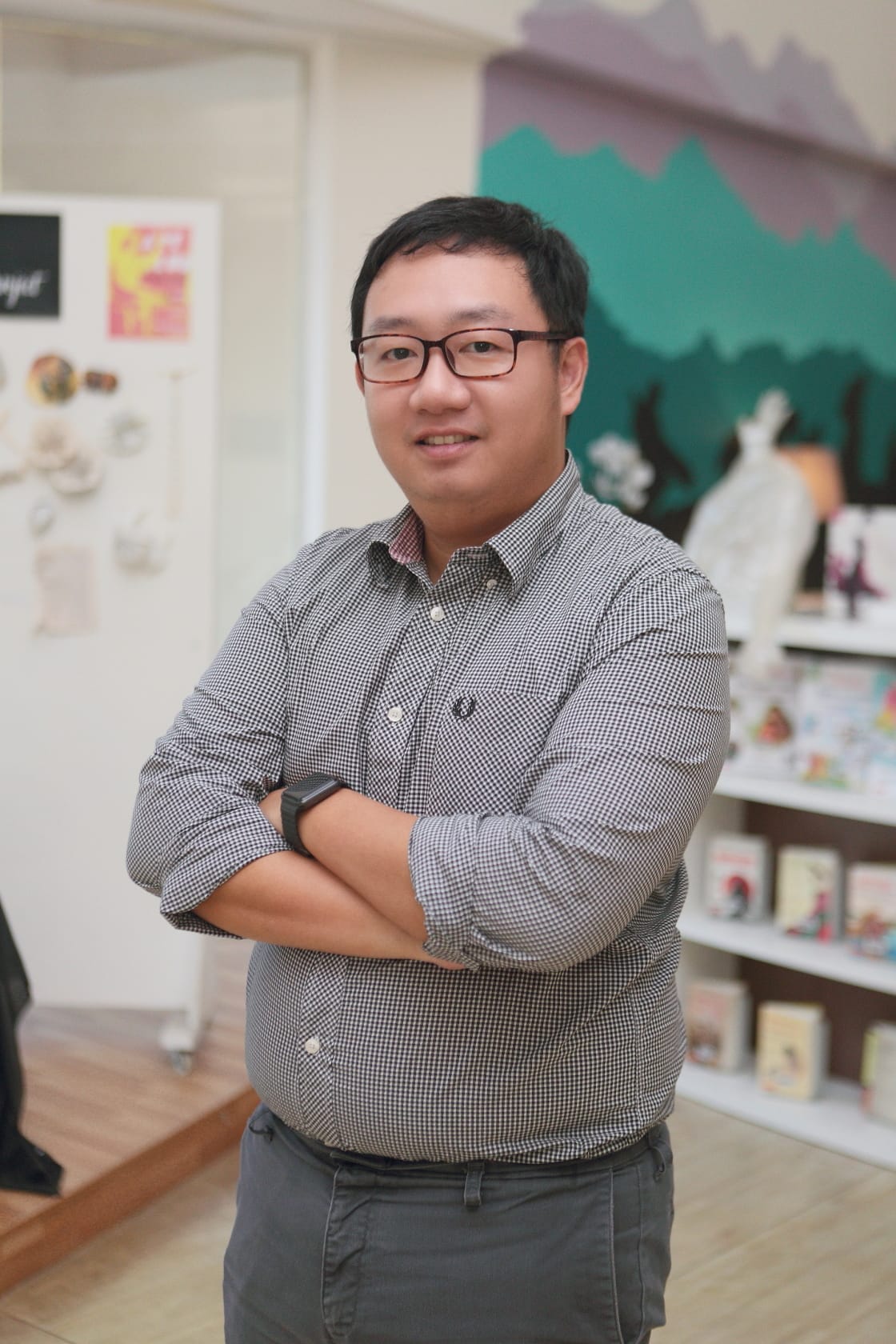 Man with glasses and crossed arms in a checkered shirt smiling, standing in a bright room with shelves and wall art.