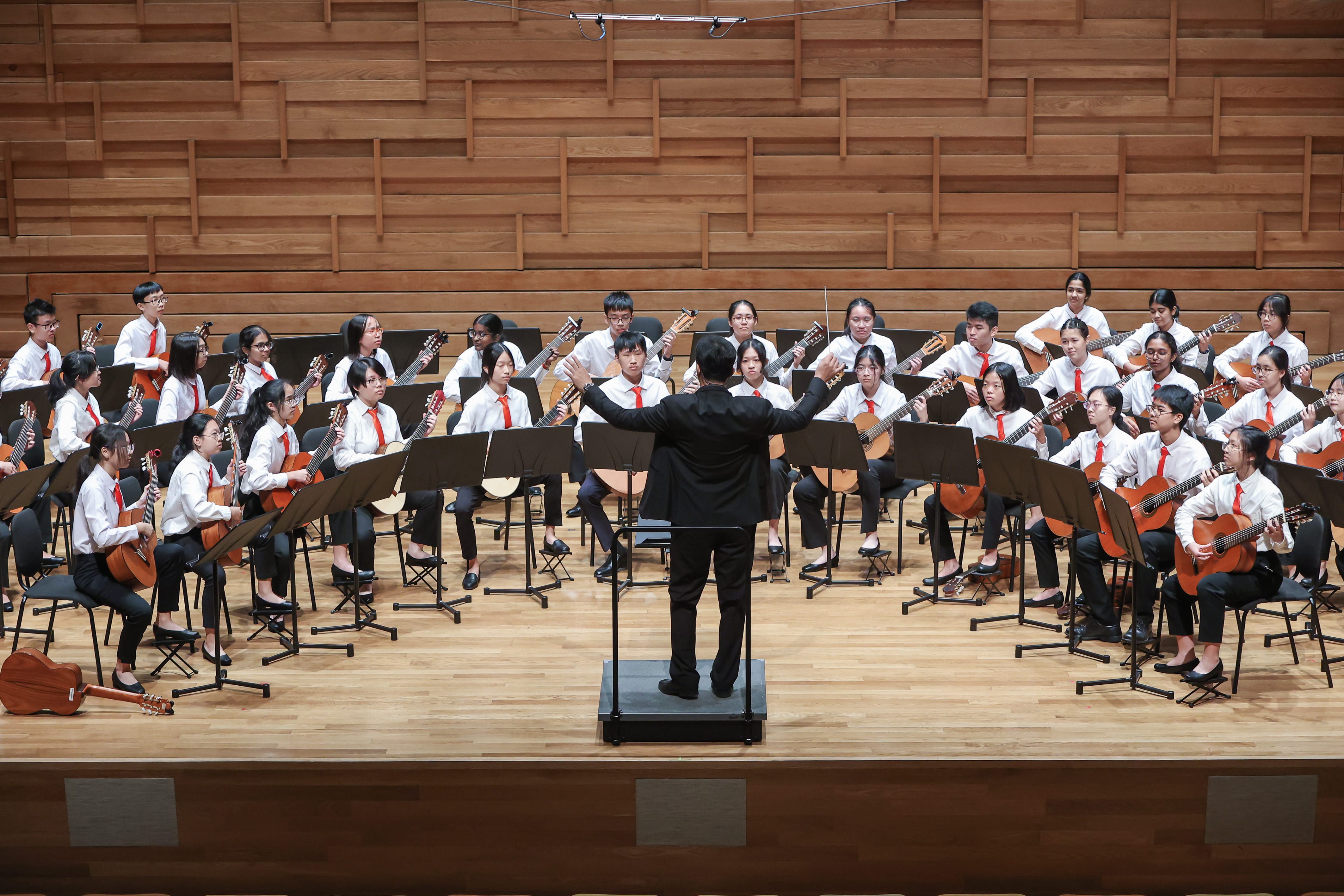 A conductor in black leads a large youth guitar ensemble in white shirts and red ties on a wooden stage.