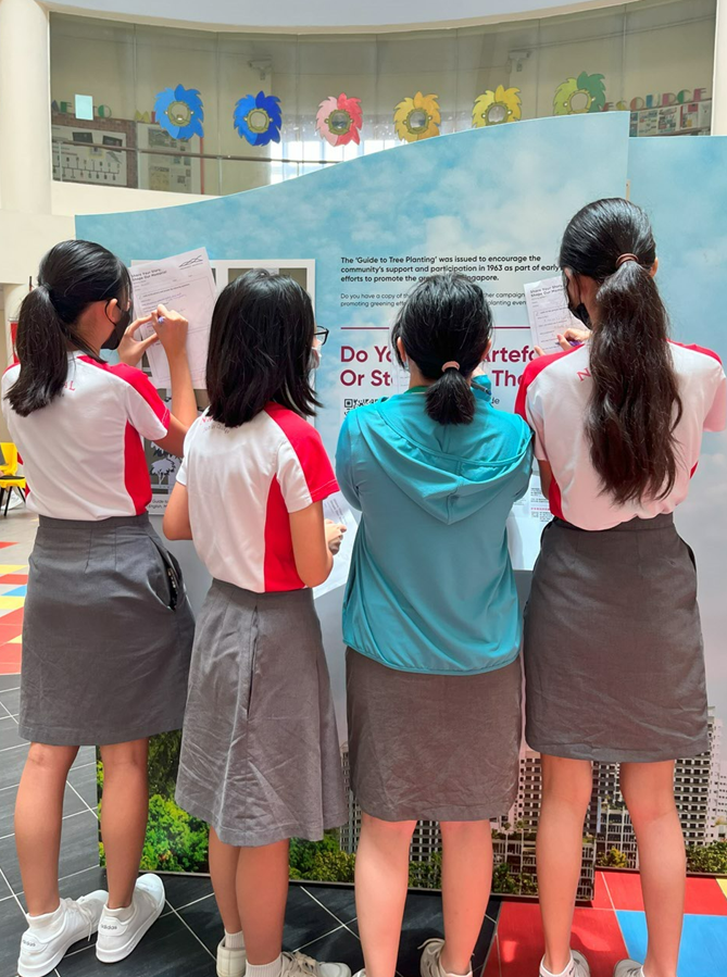 Four girls in school uniforms stand with their backs turned, reading an informational display board indoors.