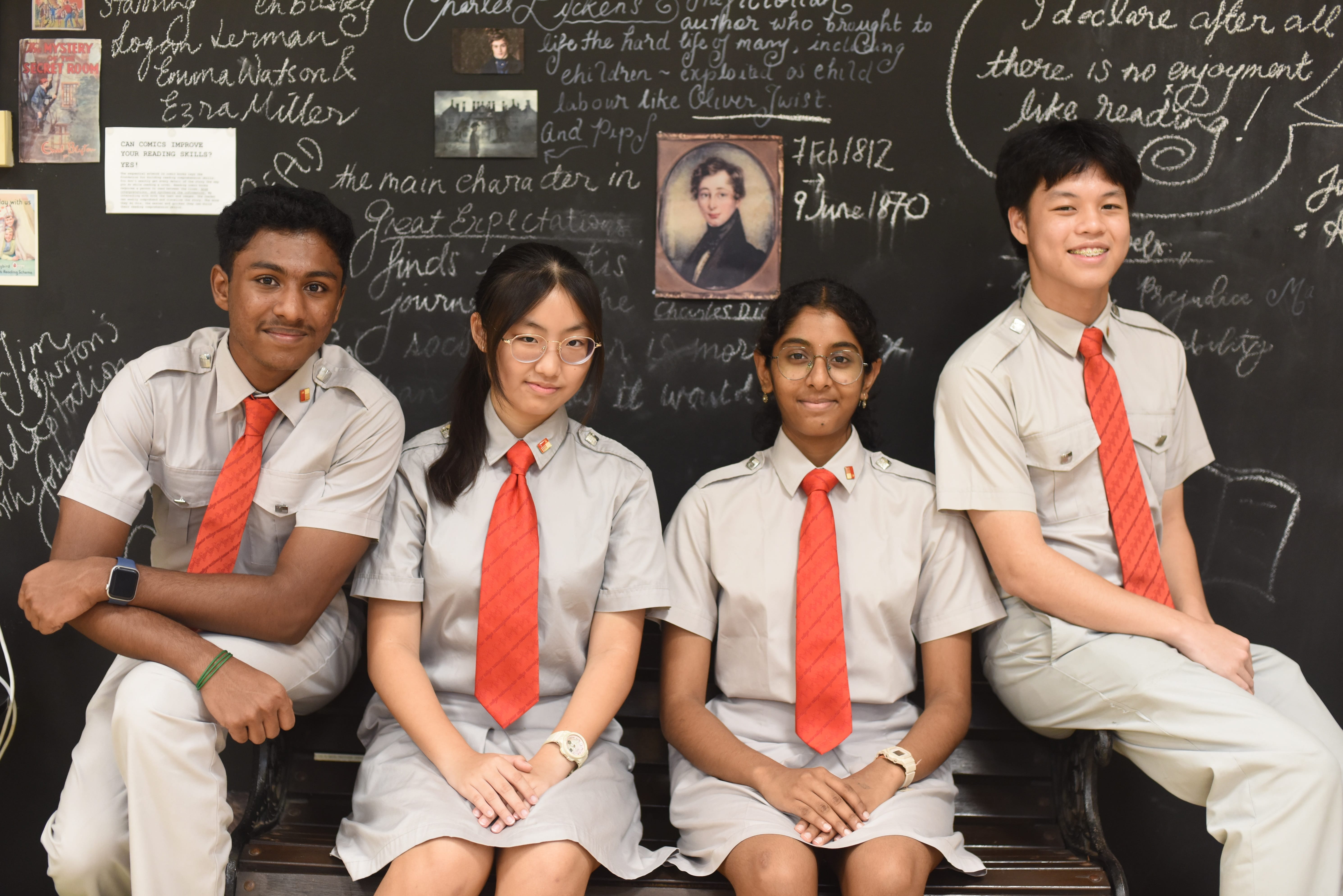 Four students in gray uniforms with red ties sit on a bench before a chalkboard with writing and photos.