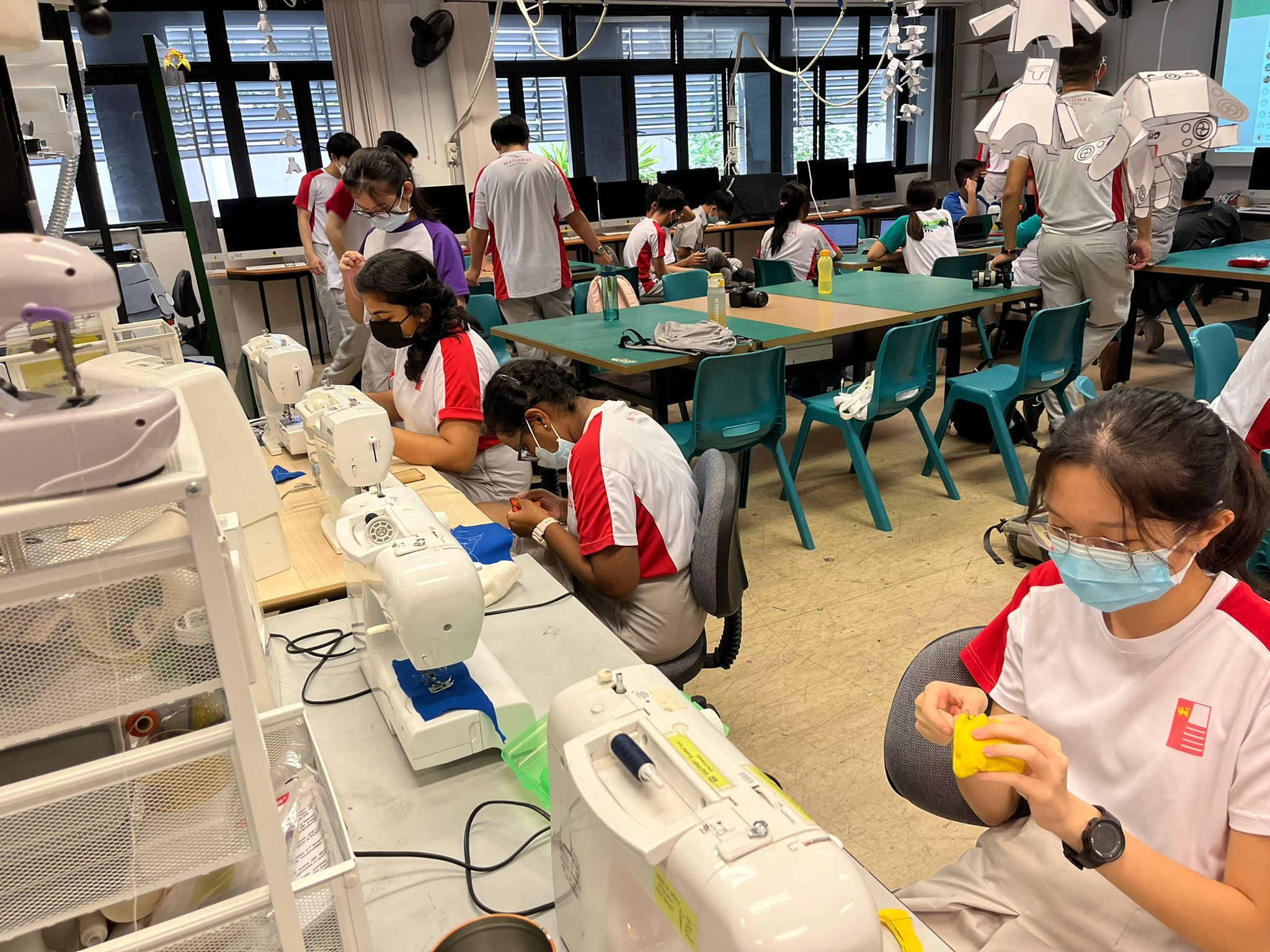 Students in masks sew at machines in a classroom while others work at tables; one holds yellow fabric.