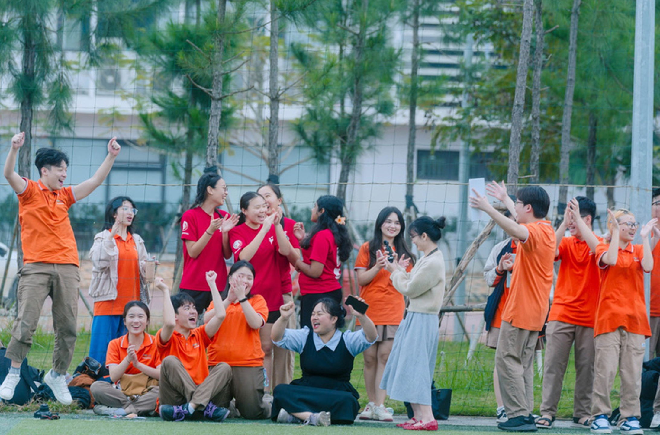 Group in orange and red shirts cheering and clapping on a grassy field near a net and trees