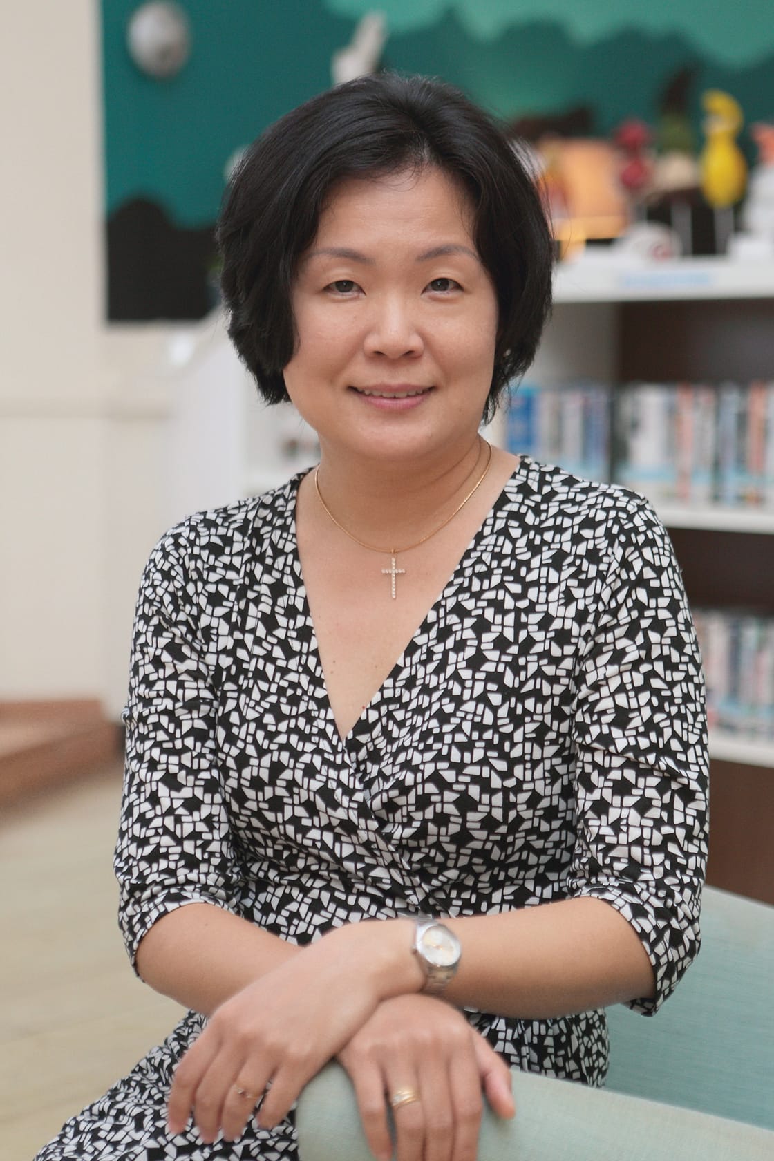 A smiling middle-aged woman in a black and white patterned top, cross necklace, and watch, seated in a library.
