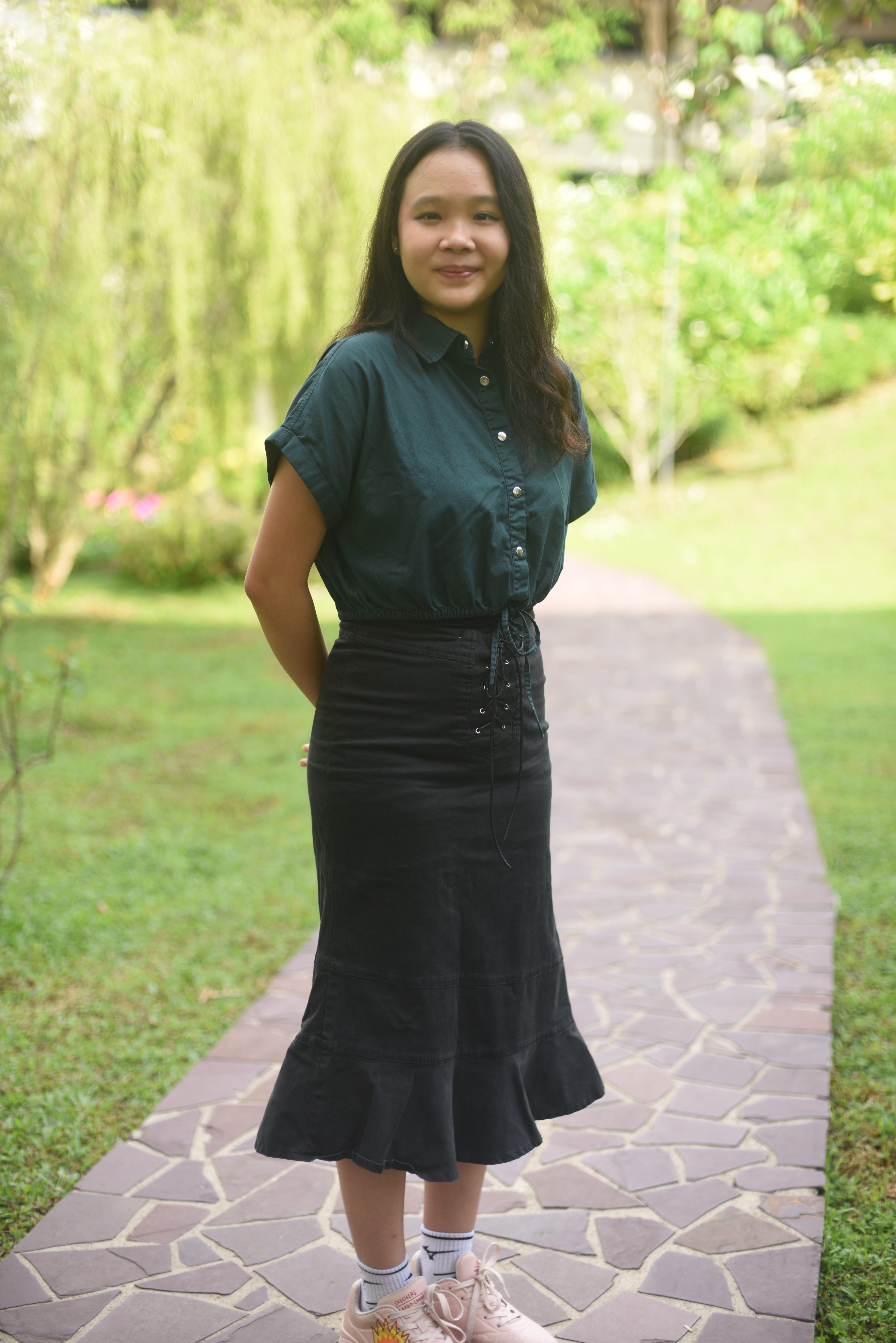 Person in green button-up shirt and black skirt standing on a stone path in a leafy park.