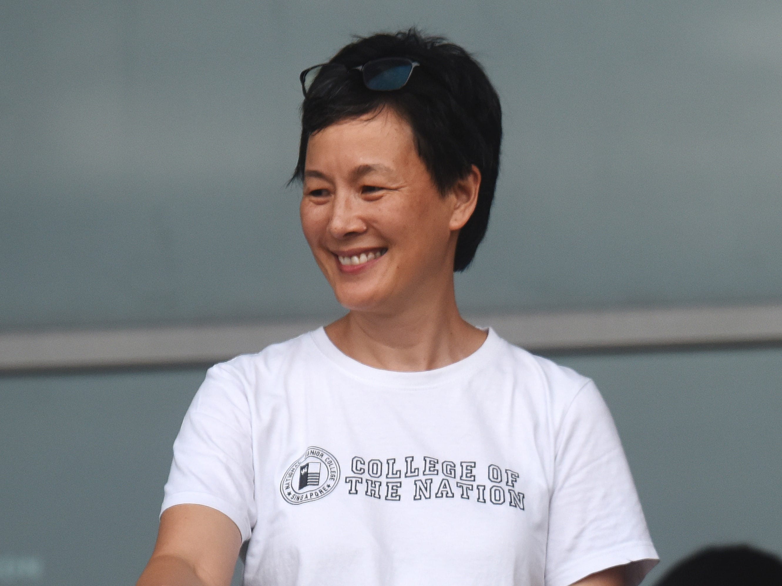 A smiling woman with short black hair and sunglasses on her head, wearing a white College of the Nation t-shirt.