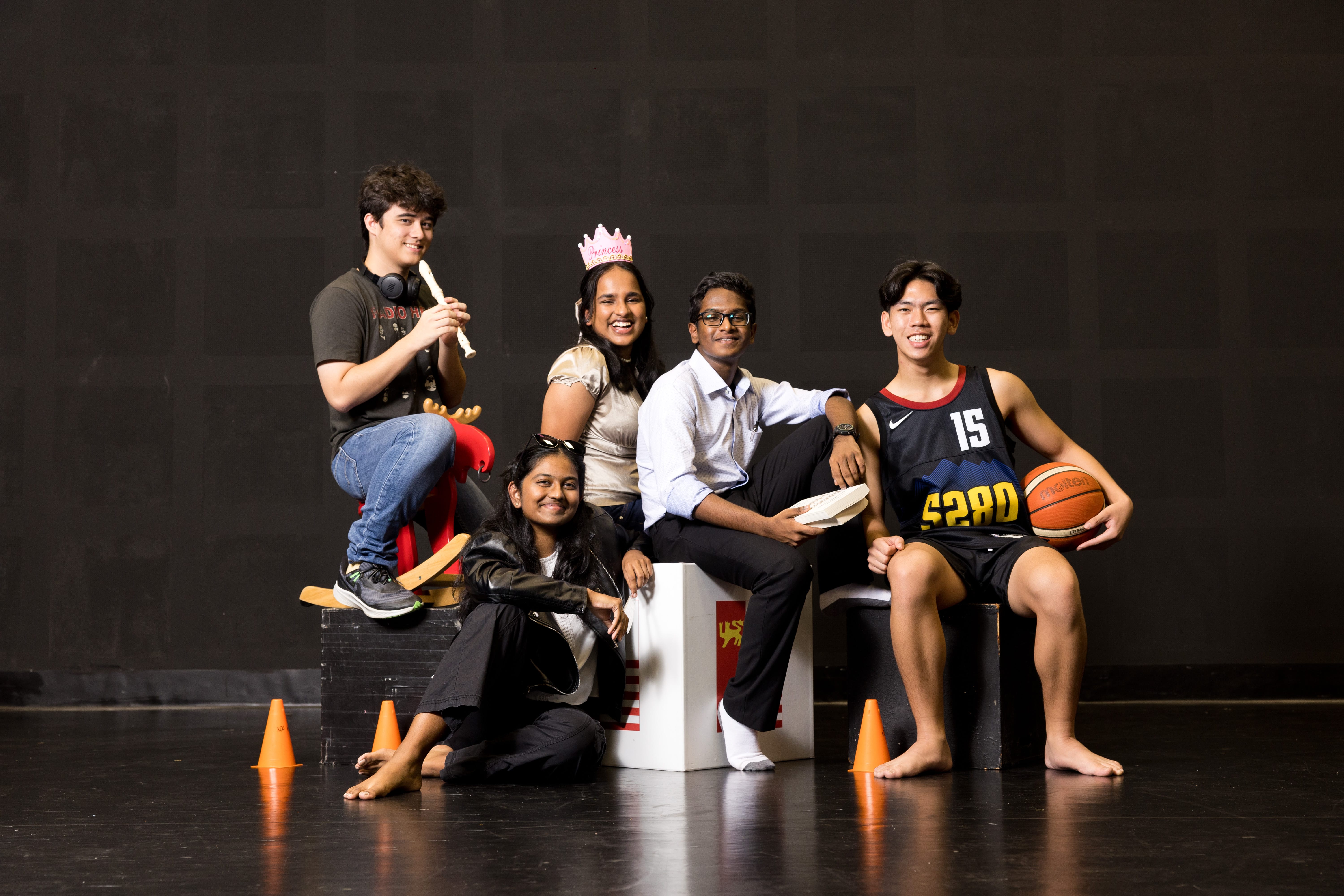 Five young adults pose on boxes; one holds flute, one wears tiara, one holds basketball; cones on floor.