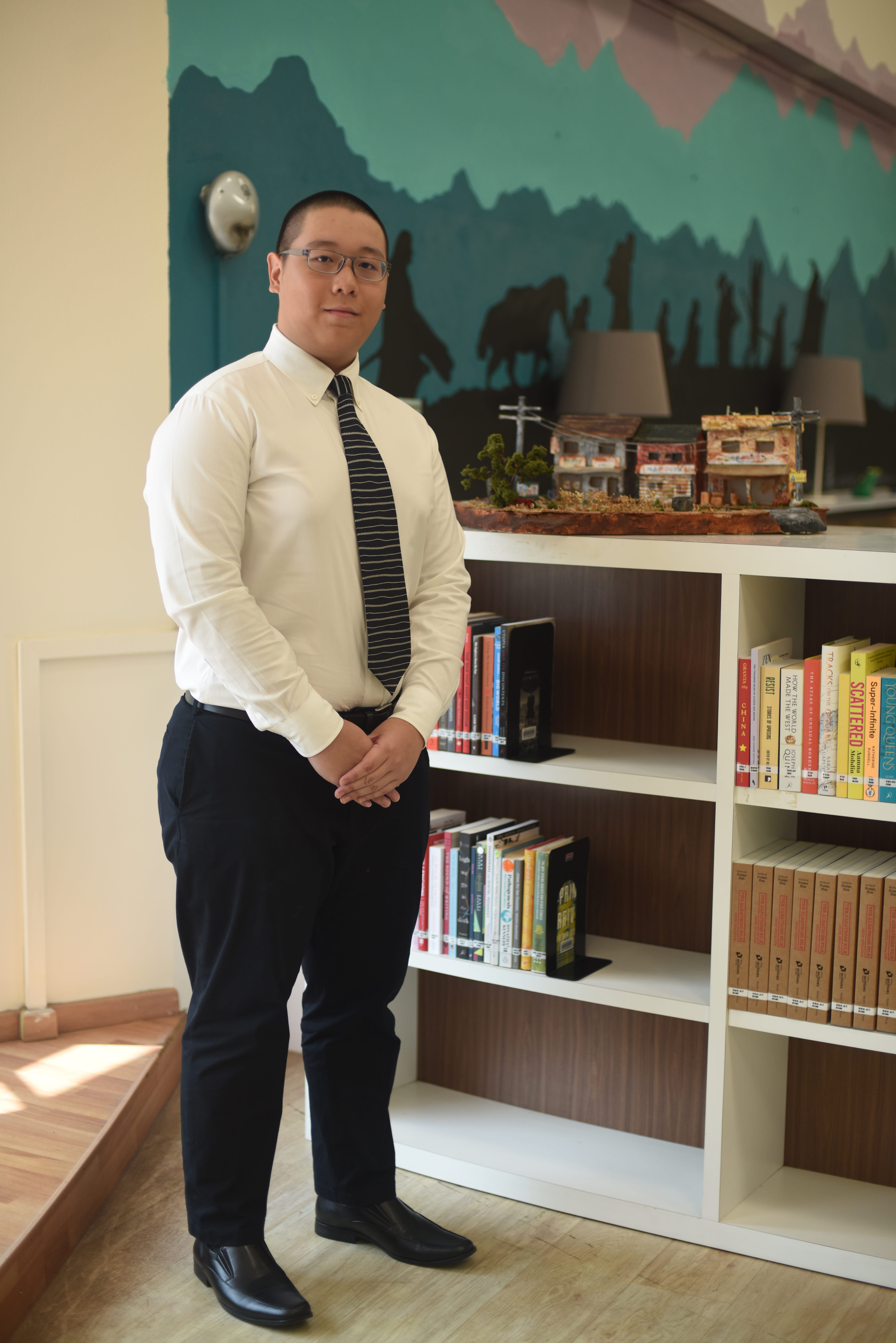 Man in white shirt and striped tie stands by a bookshelf with books and a miniature building model.