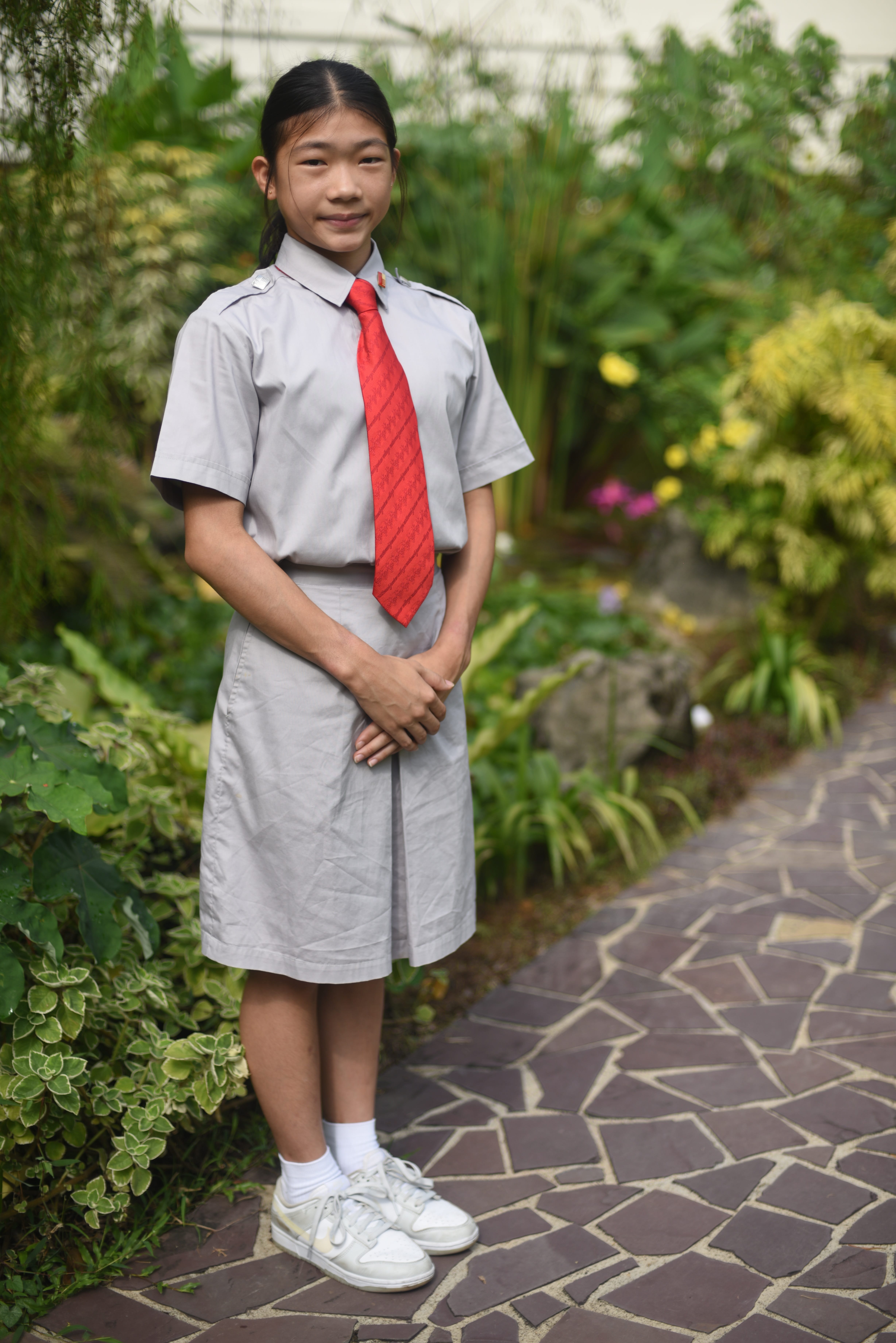 Student in gray uniform with red tie stands on stone path beside lush green plants.
