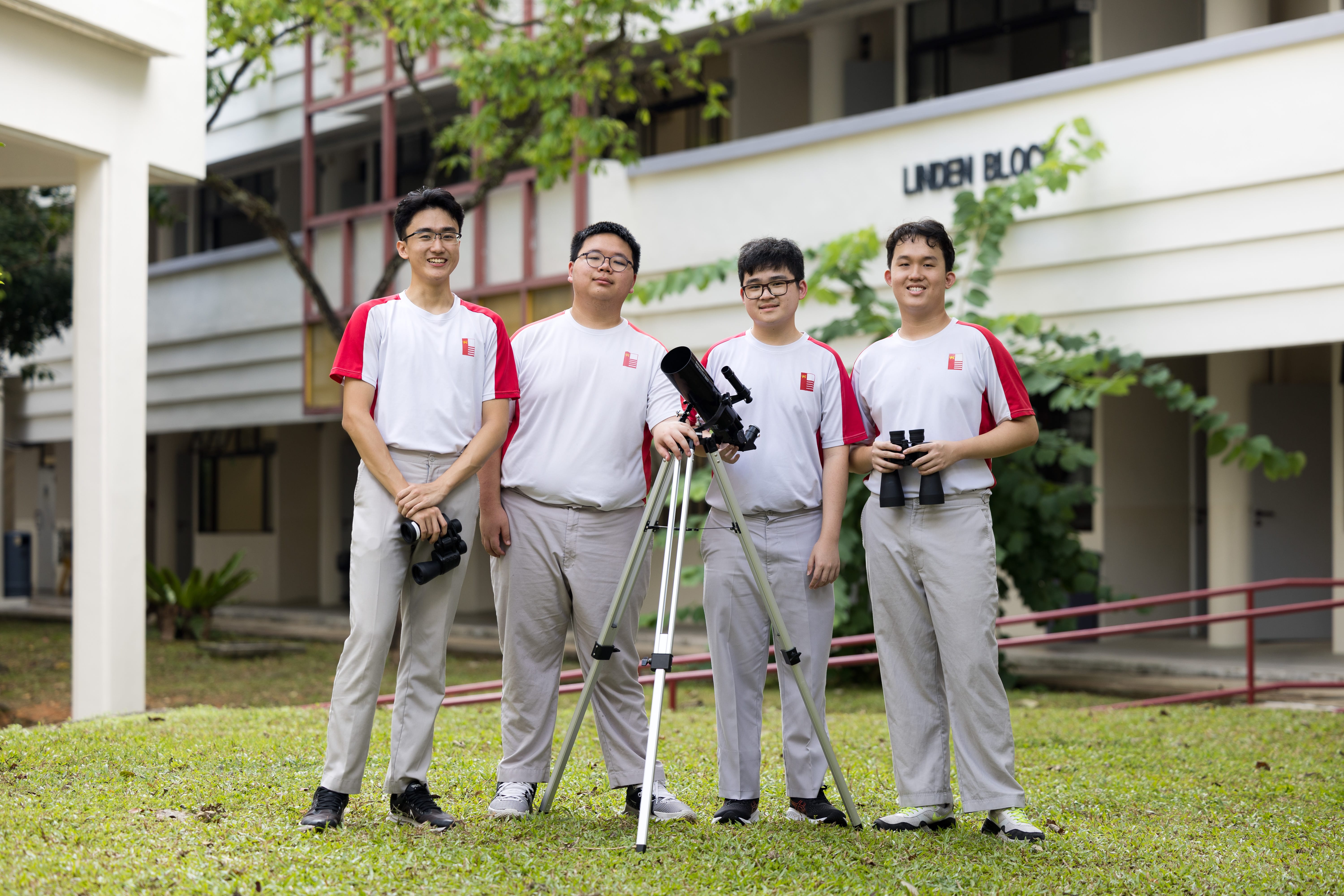 Four students in white-and-red uniforms stand on grass with a telescope on tripod and binoculars, school behind.