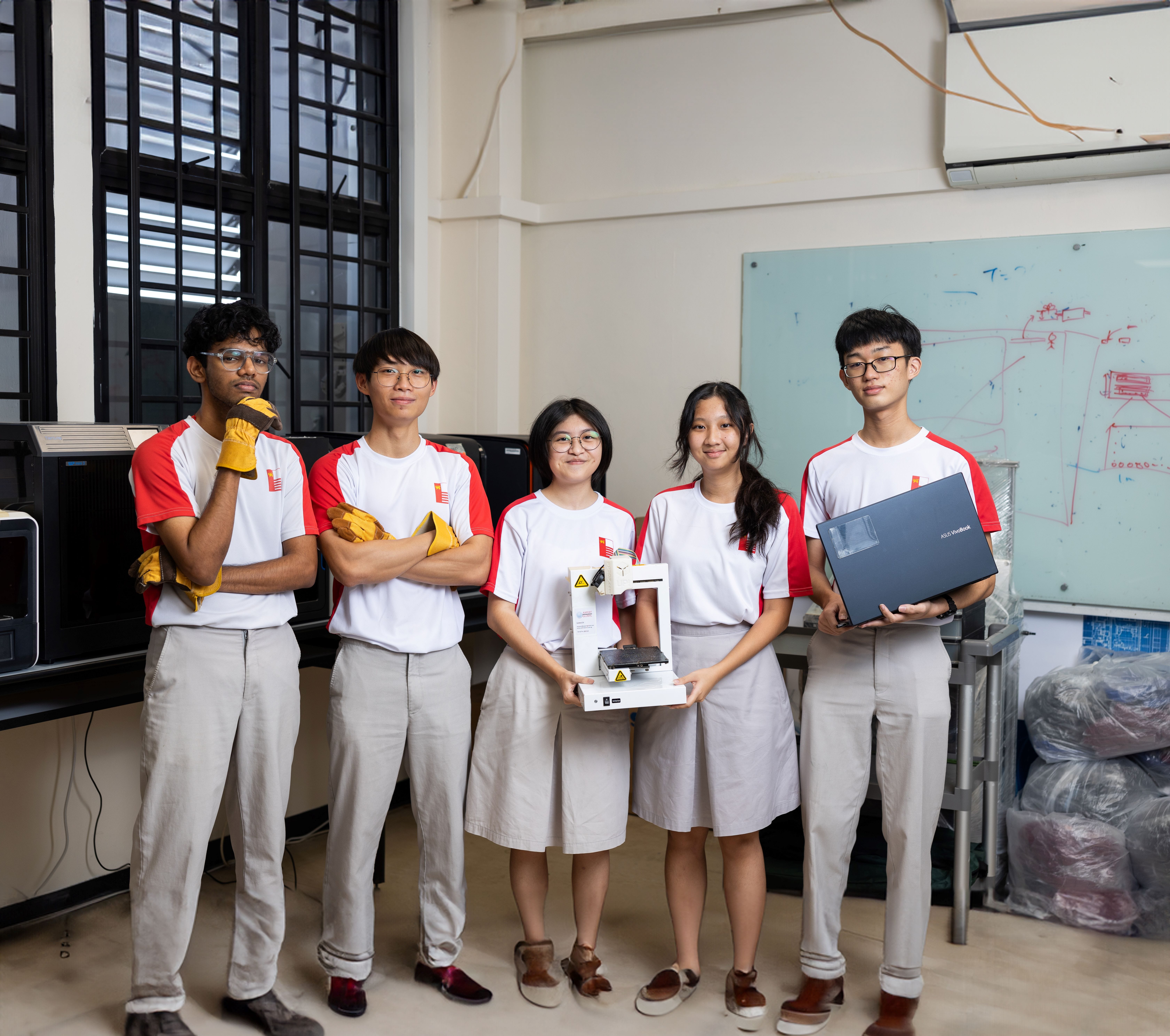 Five students in matching white and red uniforms pose in a workshop, holding a small machine and a laptop.