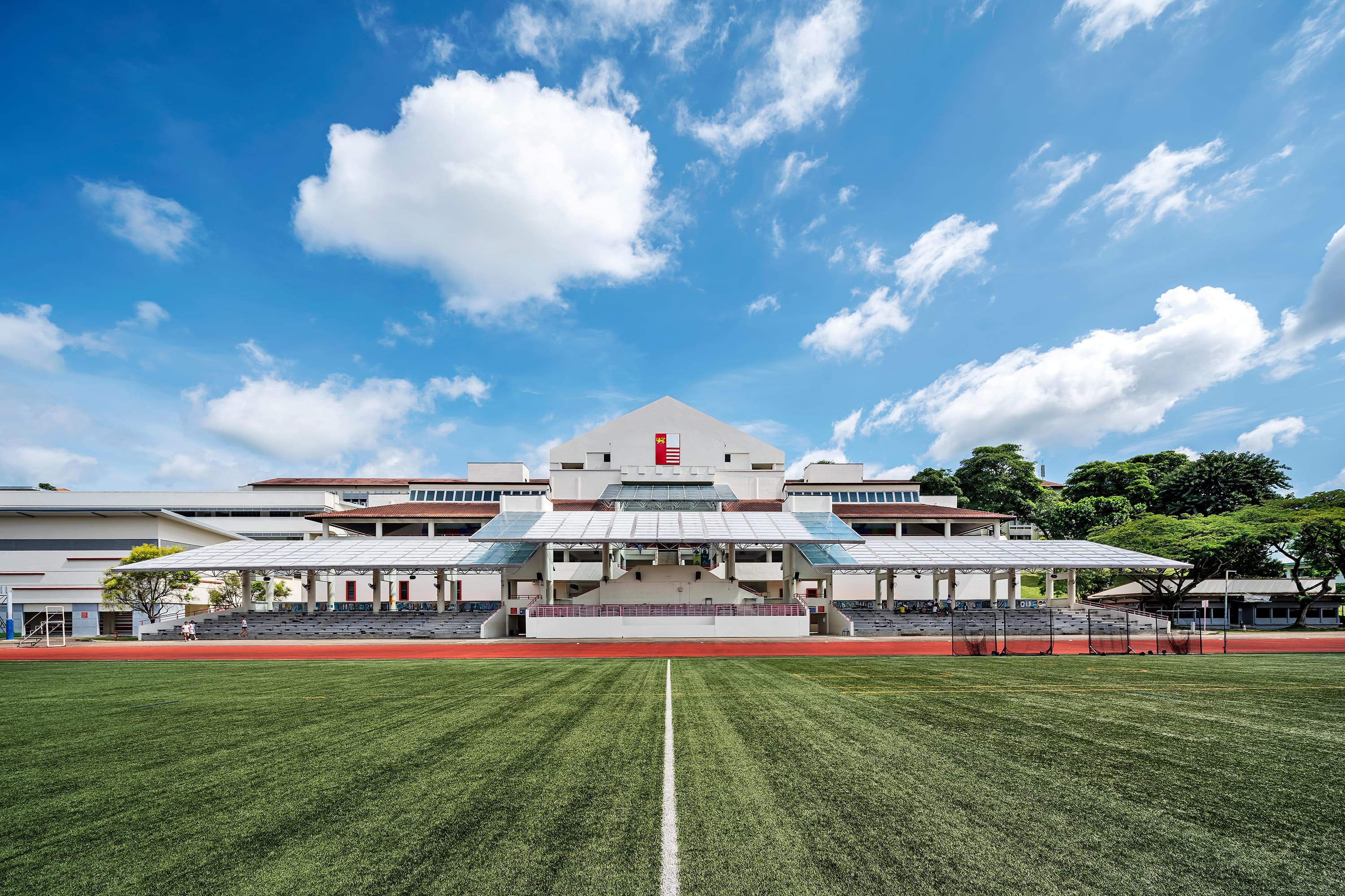 Sports field facing a stadium grandstand under a bright blue sky with large white clouds.