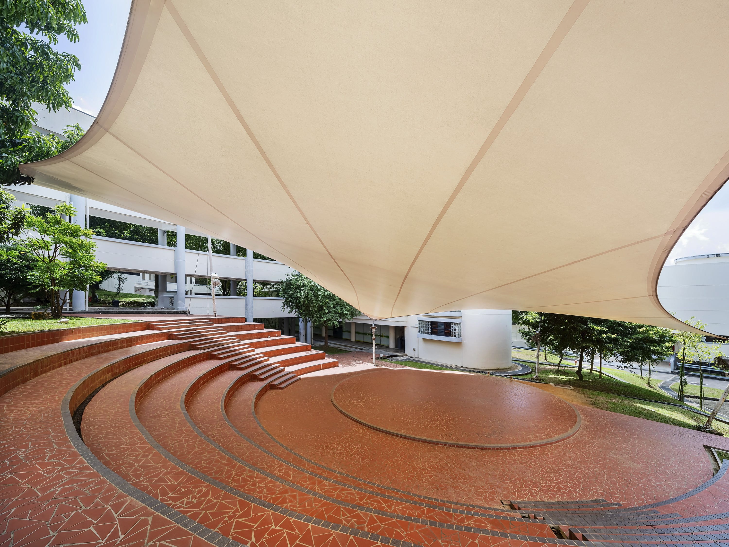 Outdoor amphitheater with red mosaic circular steps under a large cream tensile fabric canopy, surrounded by white buildings and trees.