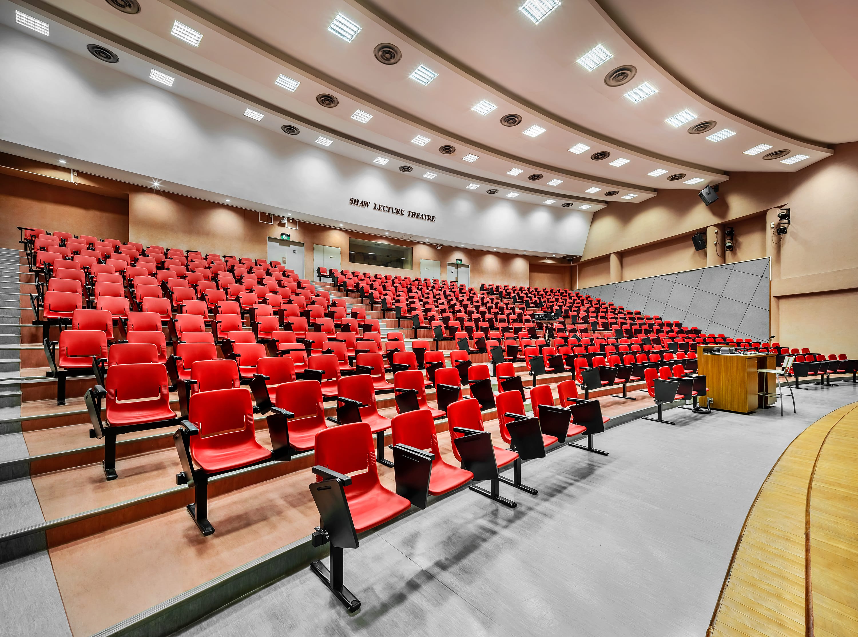 Empty lecture hall with tiered rows of red seats, bright ceiling lights, and a wooden lectern at front right.