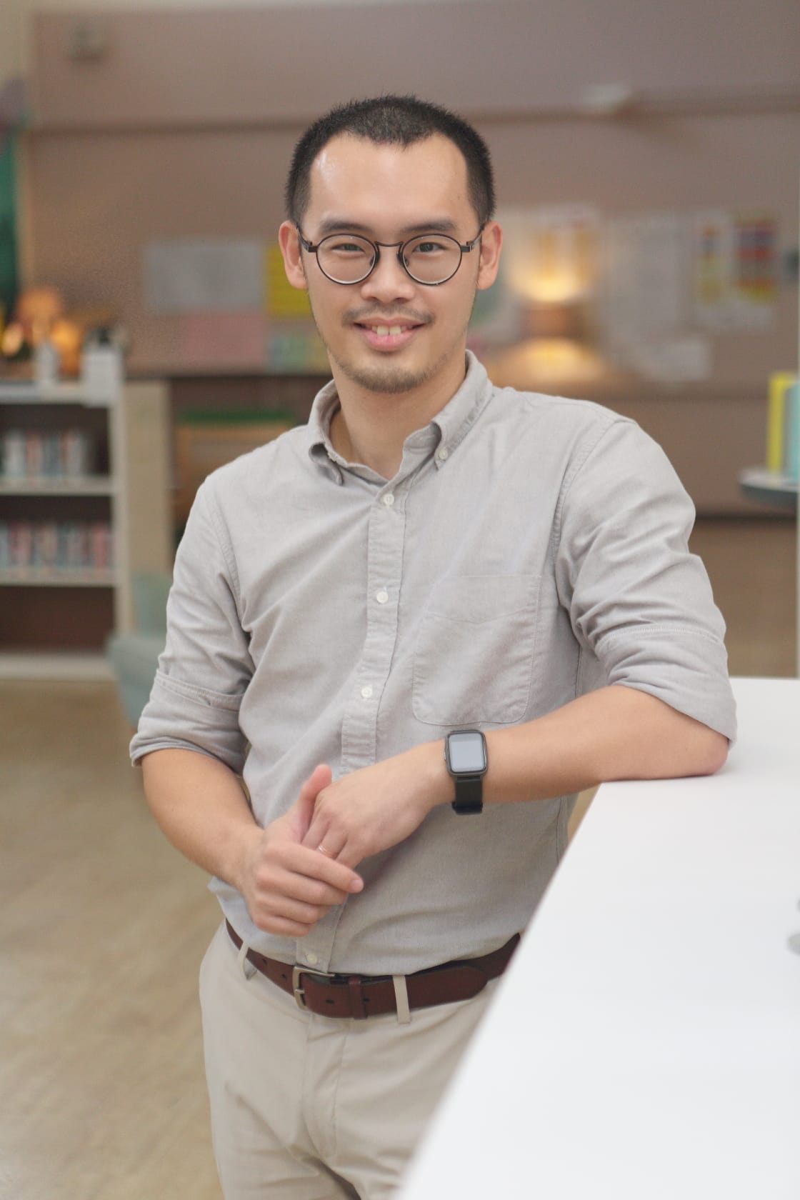 Young man with round glasses and a gray shirt smiling, leaning on a white surface in a blurred library setting.