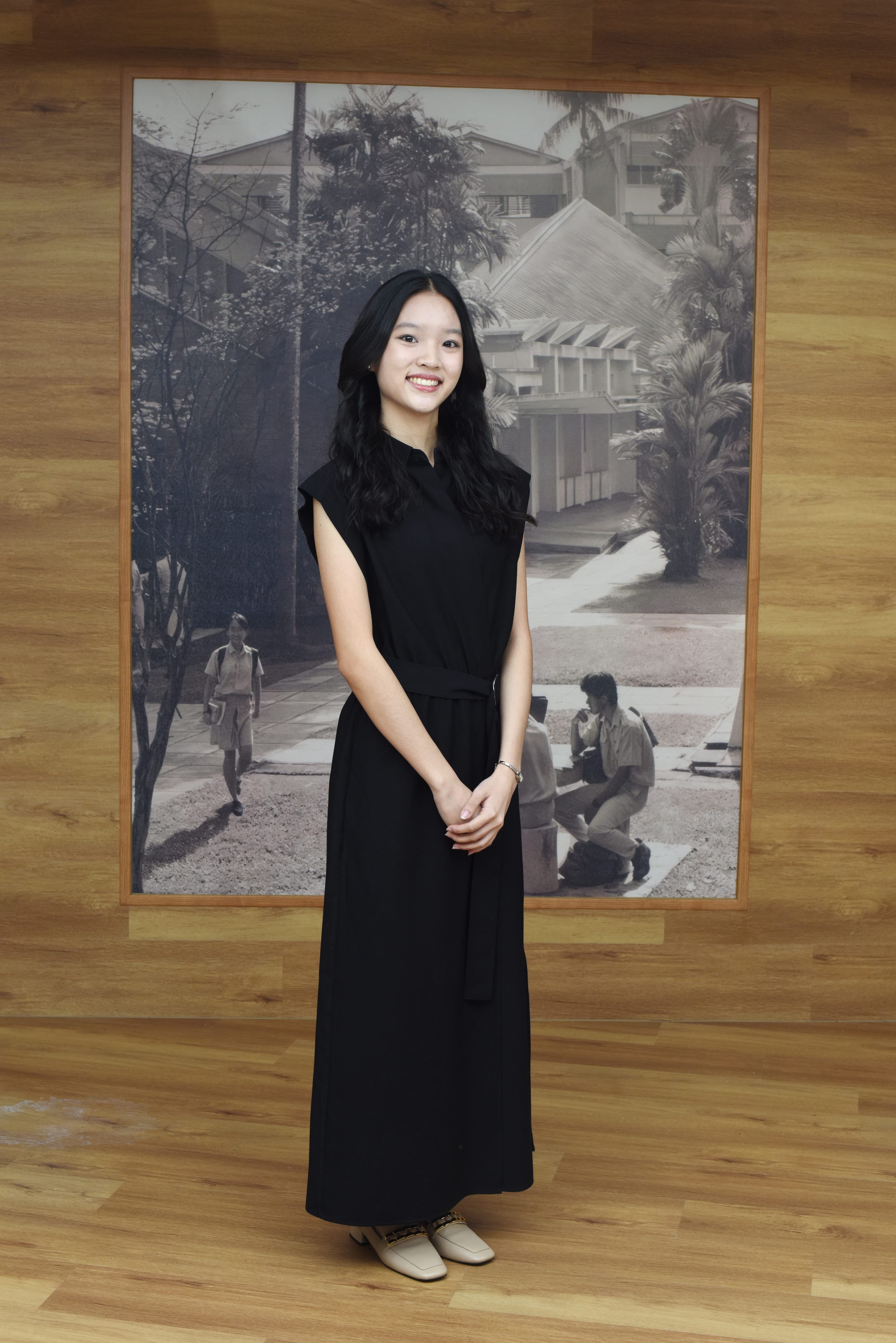 Young woman with long black hair wearing a black maxi dress, smiling and standing before a large black-and-white photo mural.