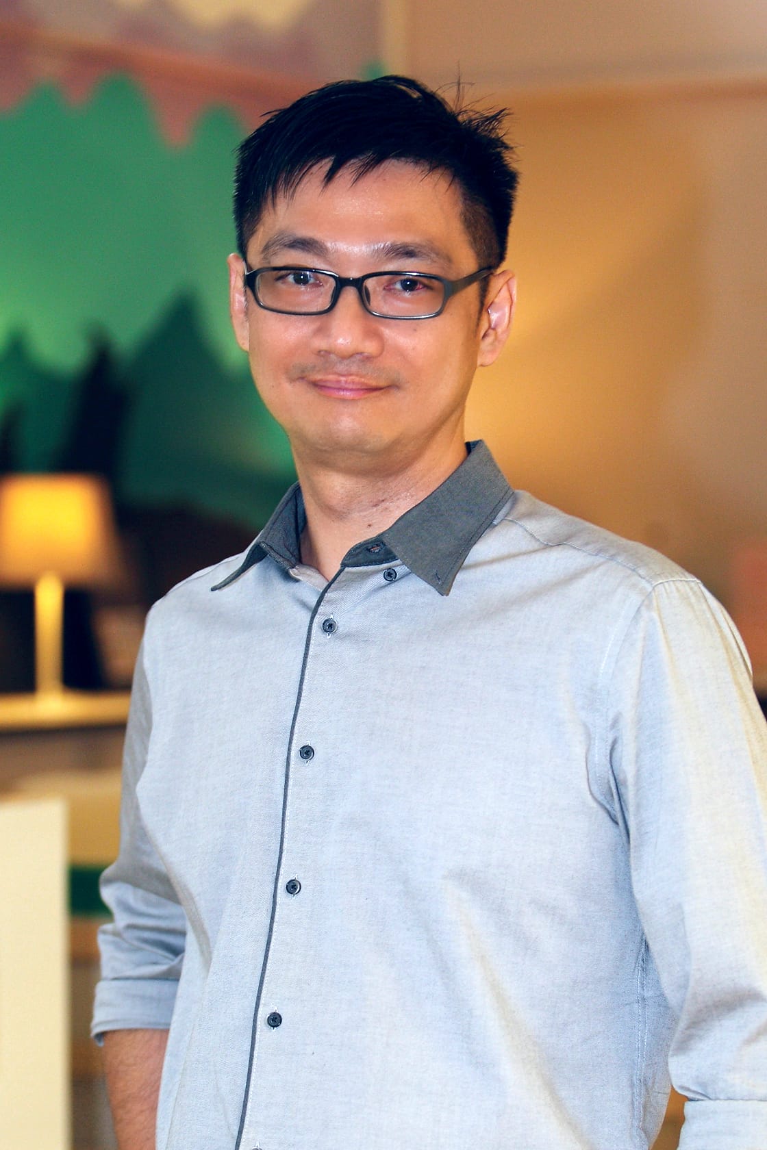 A man with black glasses and short dark hair smiles, wearing a light gray button-up shirt indoors.