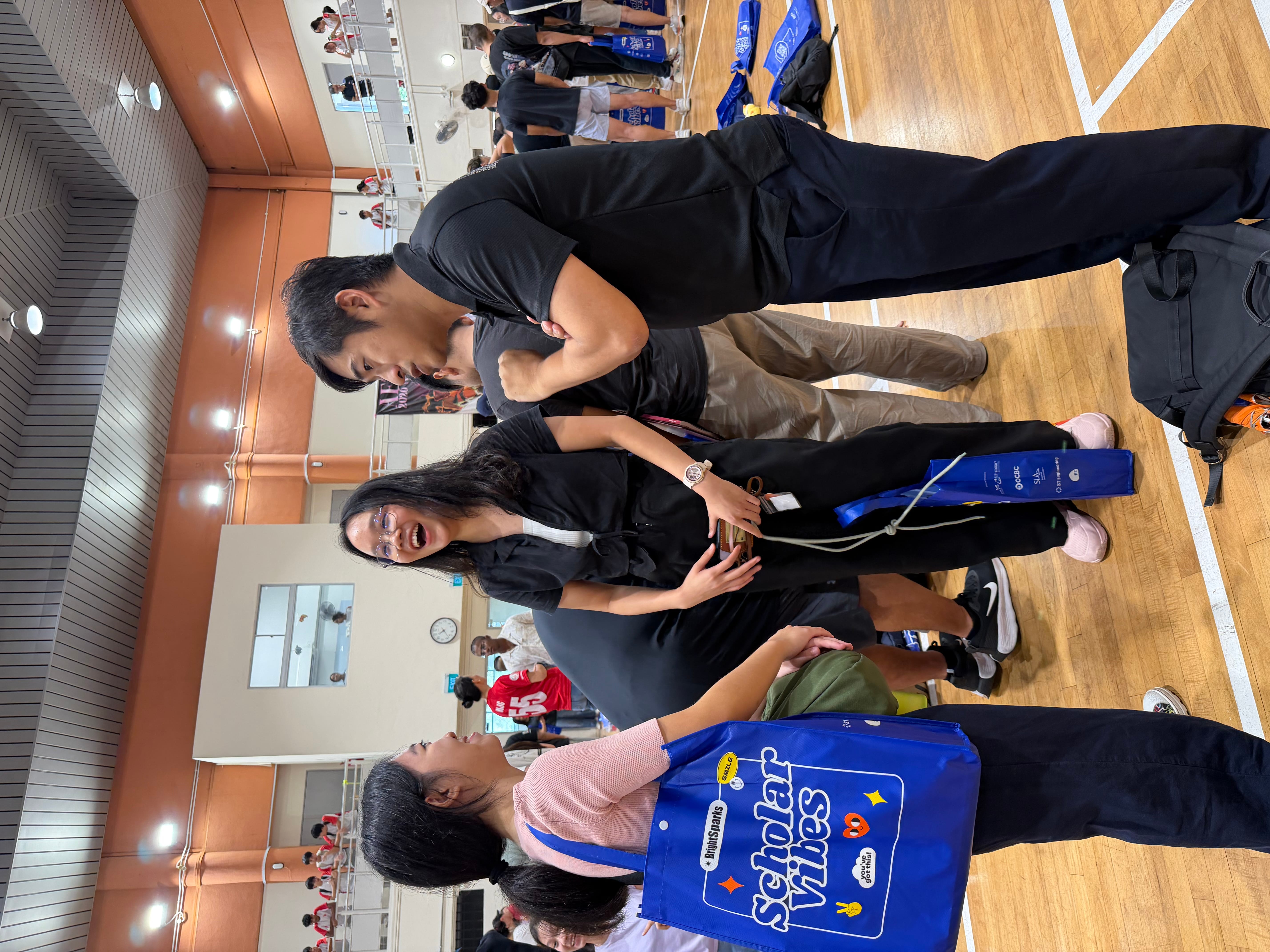 Three people chat in a gym, two holding blue tote bags, with others and bleachers in the background.
