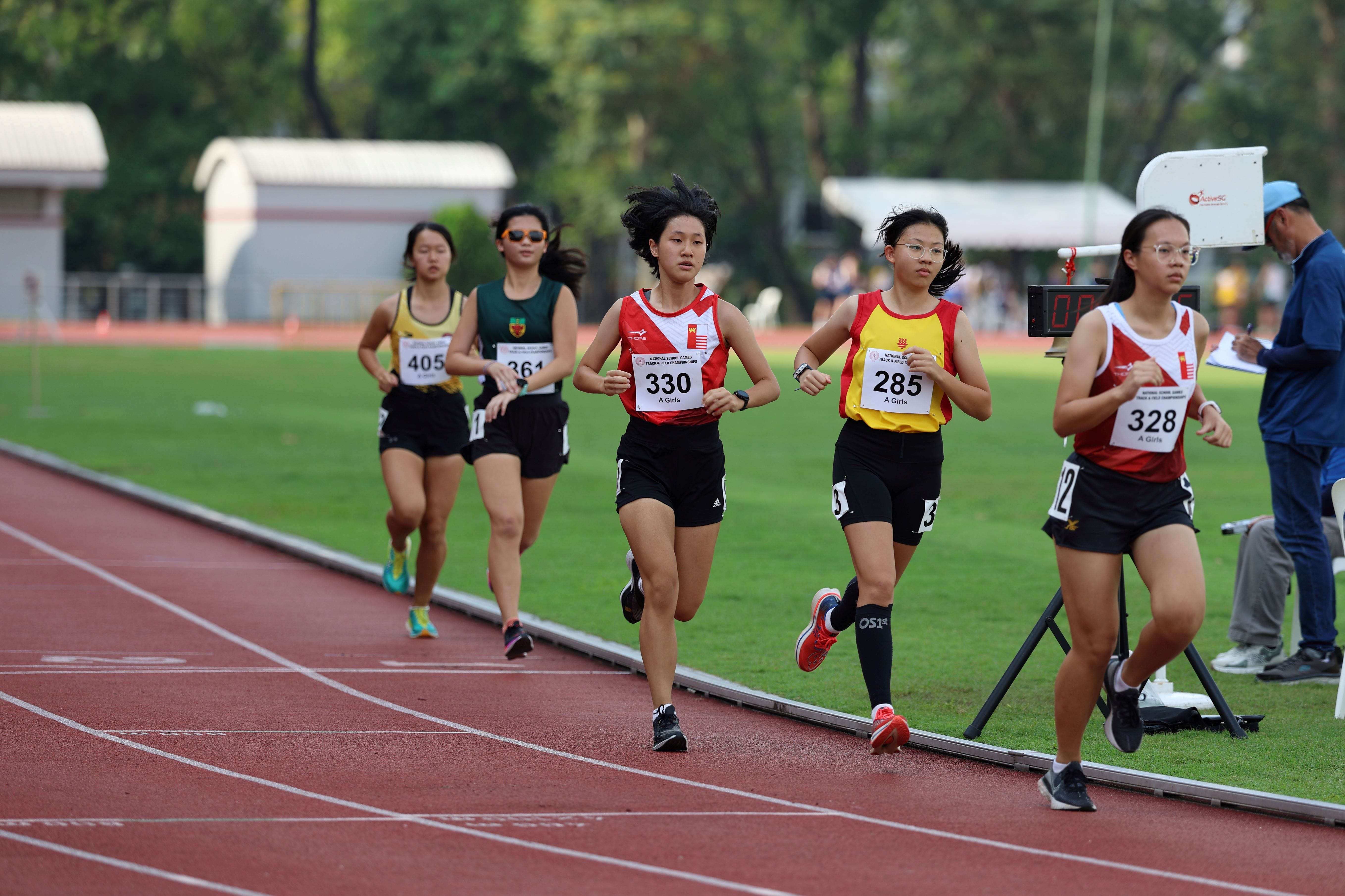 Five young female runners compete on a red track, wearing numbered bibs including 330, 285, 328, 361, and 405.