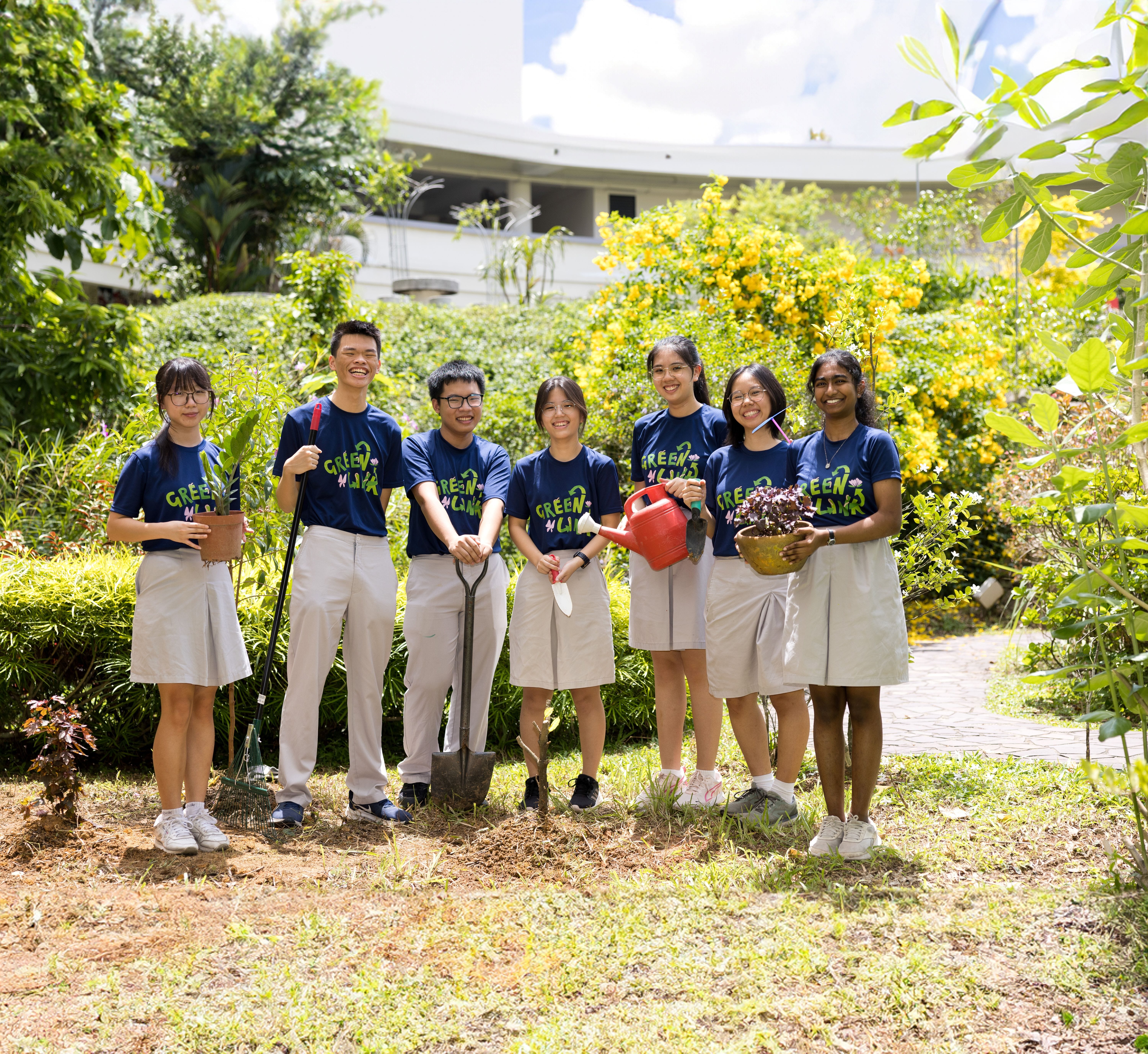 Seven students in matching shirts and uniforms pose in a garden, holding a shovel, watering can, and plants.