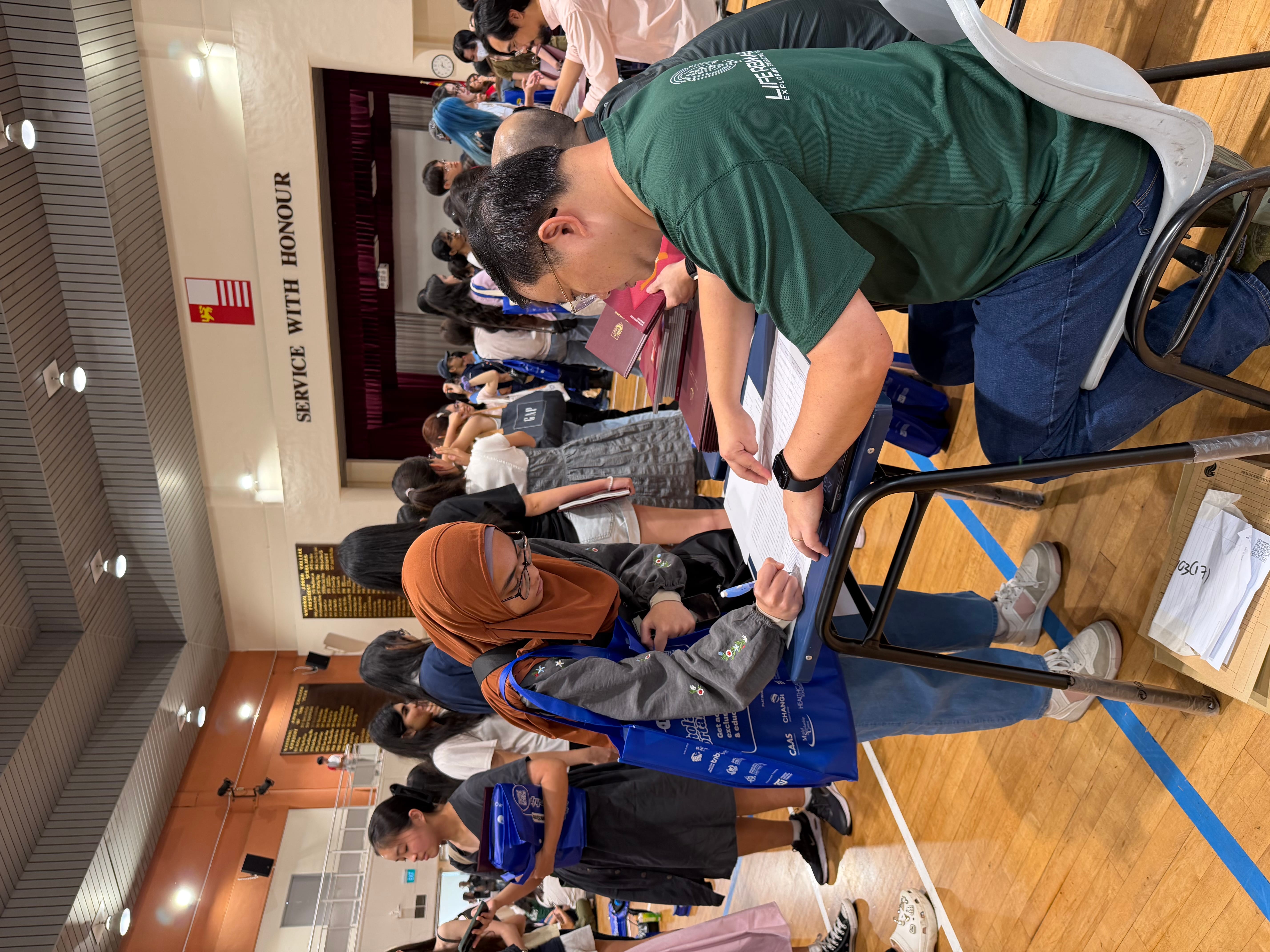 Man in green shirt signs papers at a table while a woman in orange hijab and blue tote points in a busy hall.