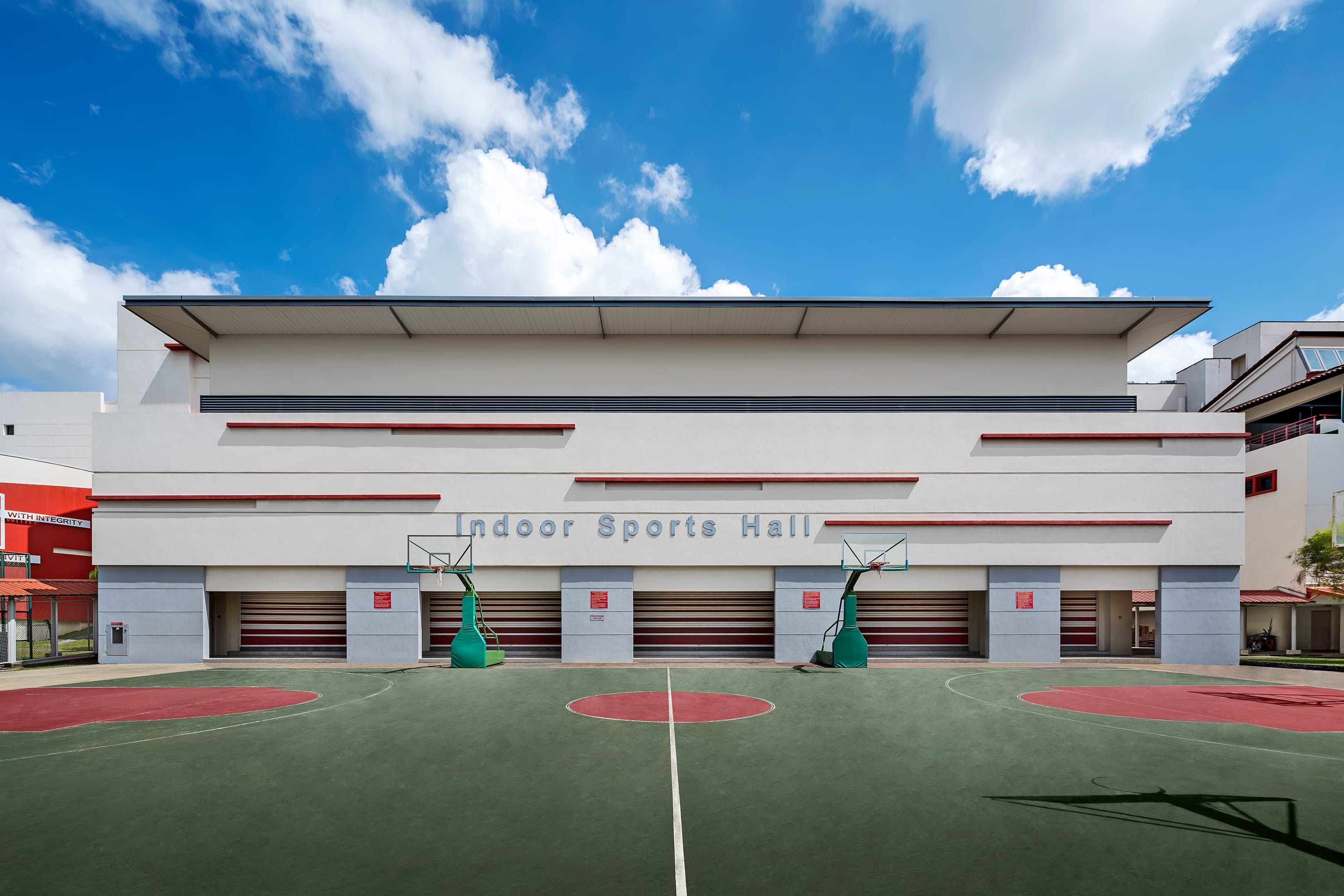 Outdoor basketball court with red markings in front of a white building labeled Indoor Sports Hall under a blue sky.