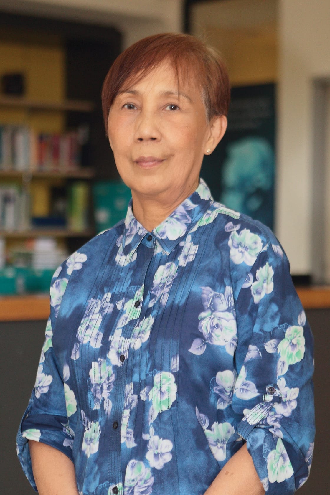Older woman with short reddish hair wearing a blue floral shirt, standing in front of a blurred bookshelf.