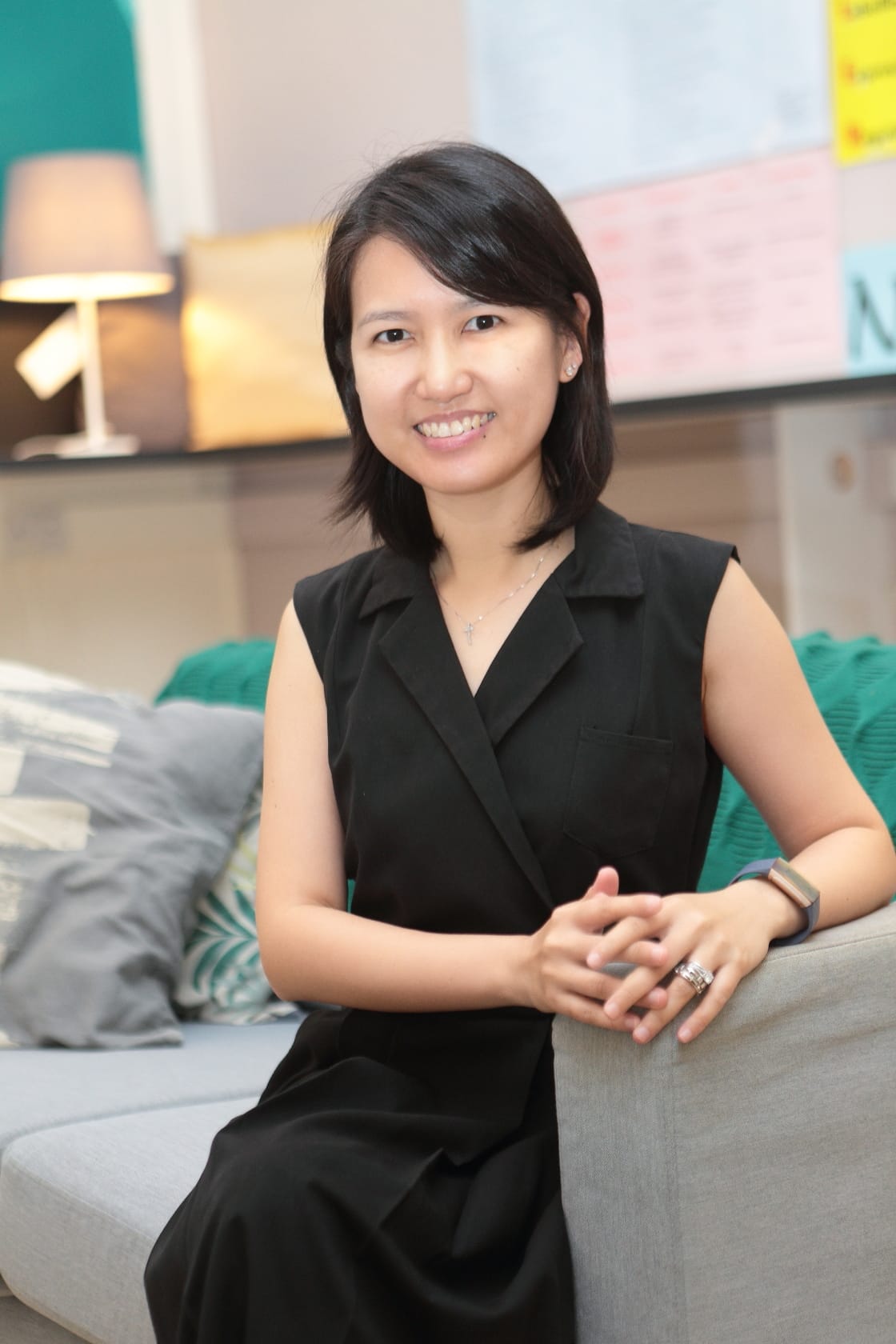 A smiling woman with short black hair in a black sleeveless dress sits on a grey sofa with hands clasped.