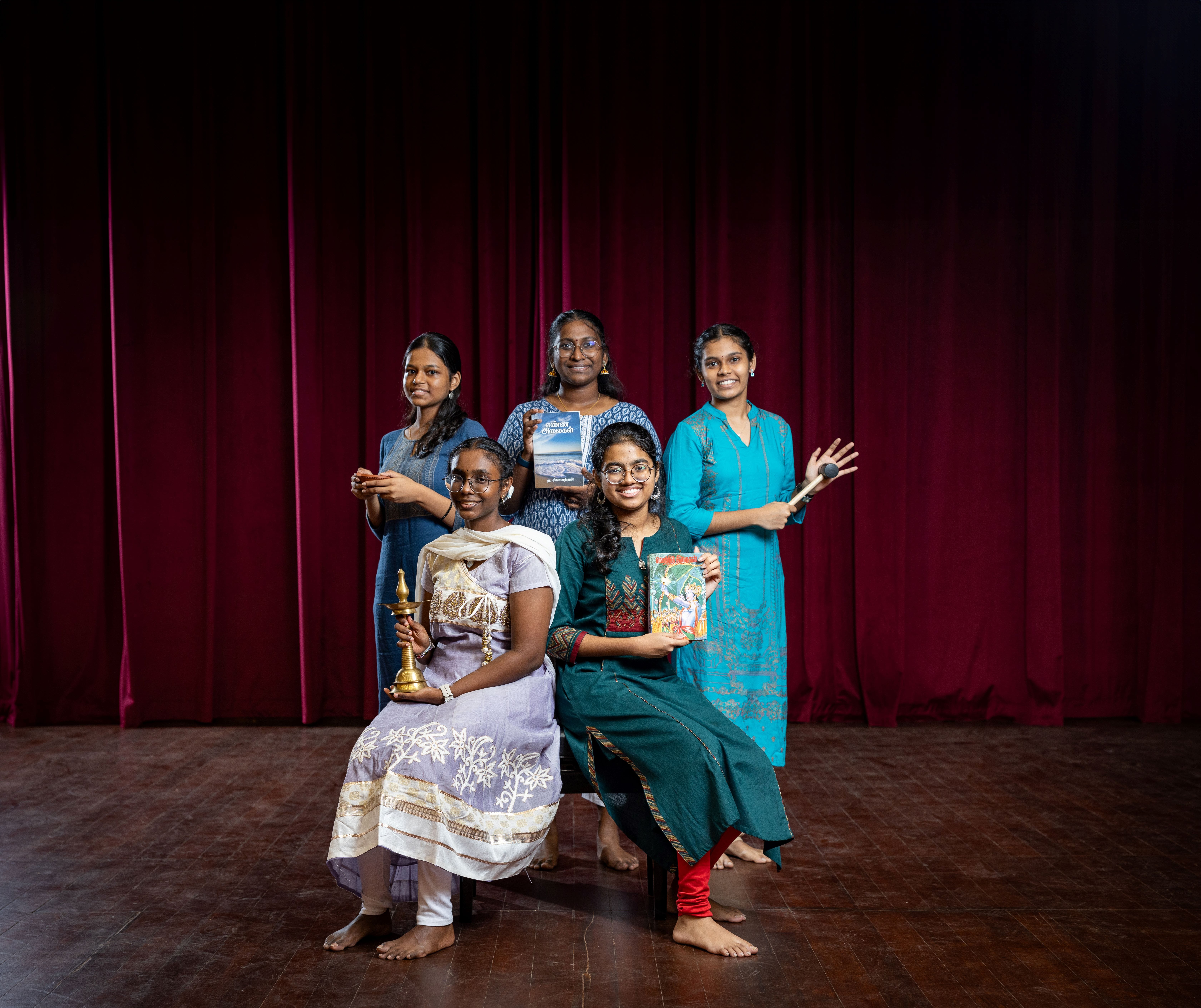 Five young women pose on a stage with a red curtain, holding books, a brass lamp, and drumsticks.