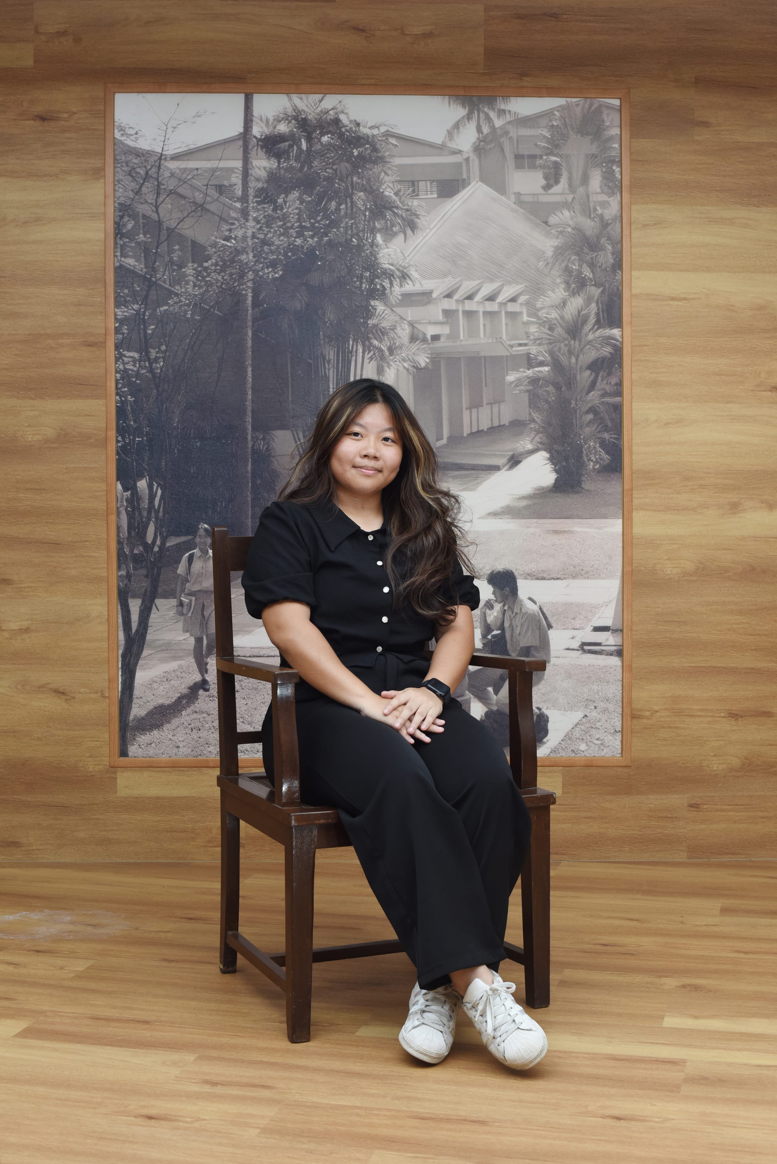 Young woman in black outfit and white sneakers sits in a wooden chair before a large black and white photo mural.