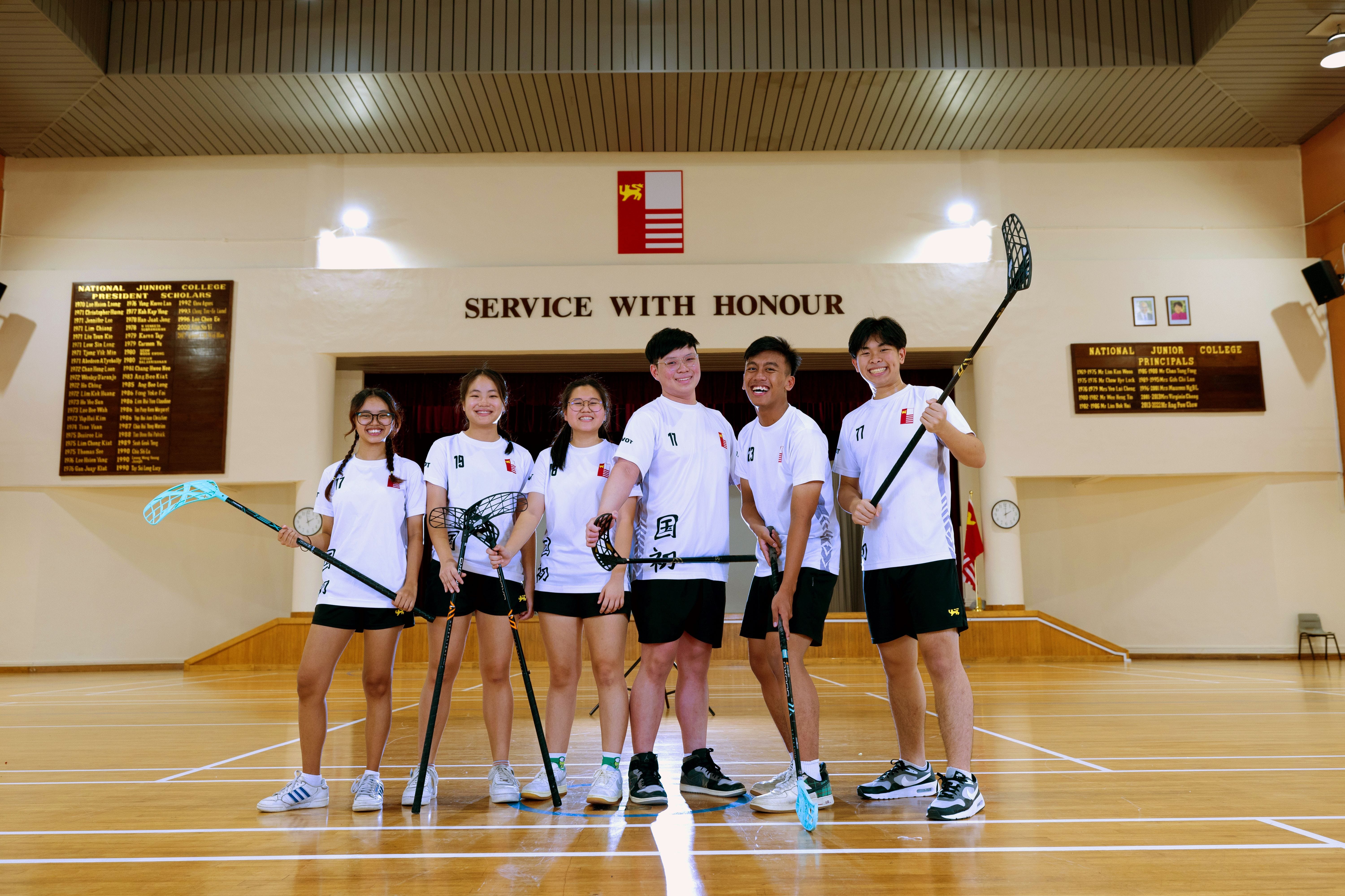 Six students in white uniforms hold floorball sticks in a school hall with a "Service With Honour" sign.