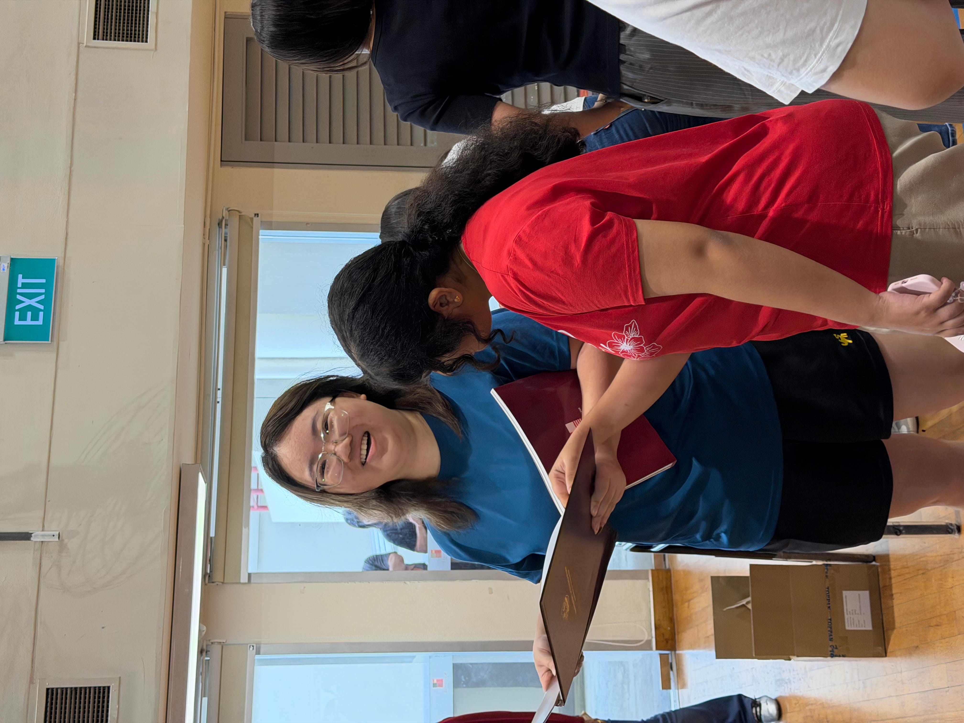 Smiling woman in blue shirt holding a dark folder, standing next to a girl in red in an indoor hall.