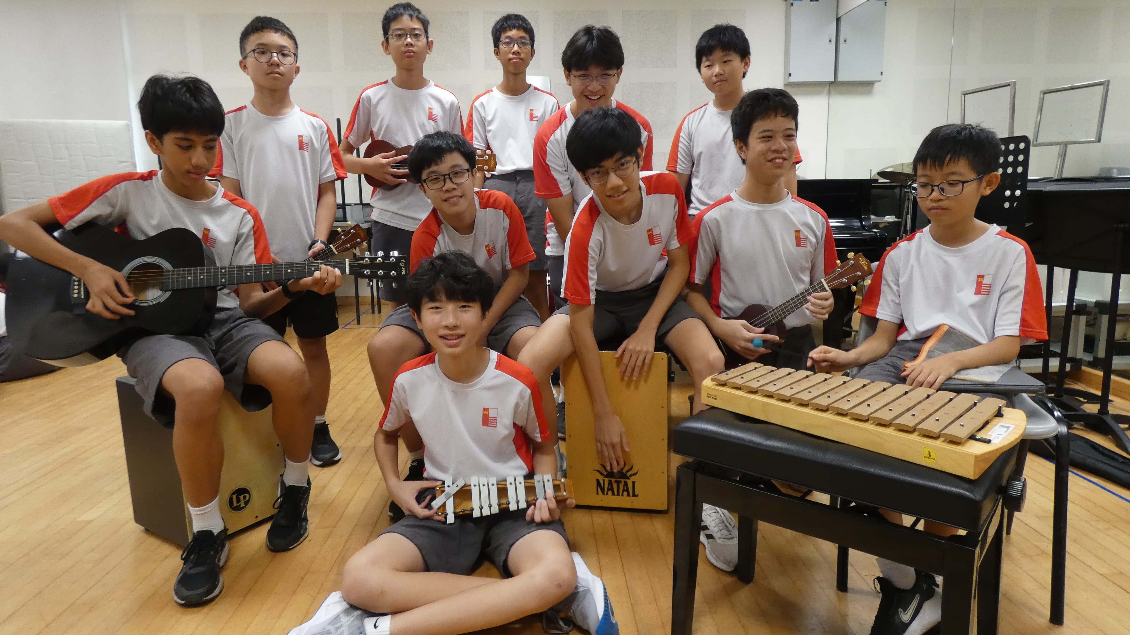 Group of students in matching uniforms in a music room, holding guitar, ukulele, xylophone, cajon.