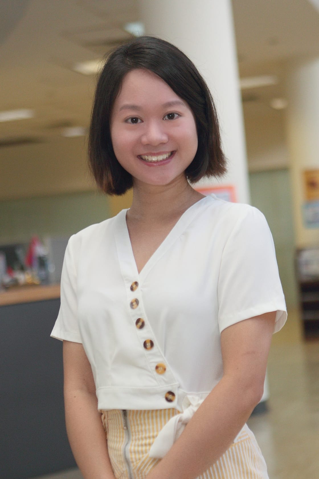 Young woman with short dark hair smiling, wearing a white button-up top and yellow striped pants indoors.