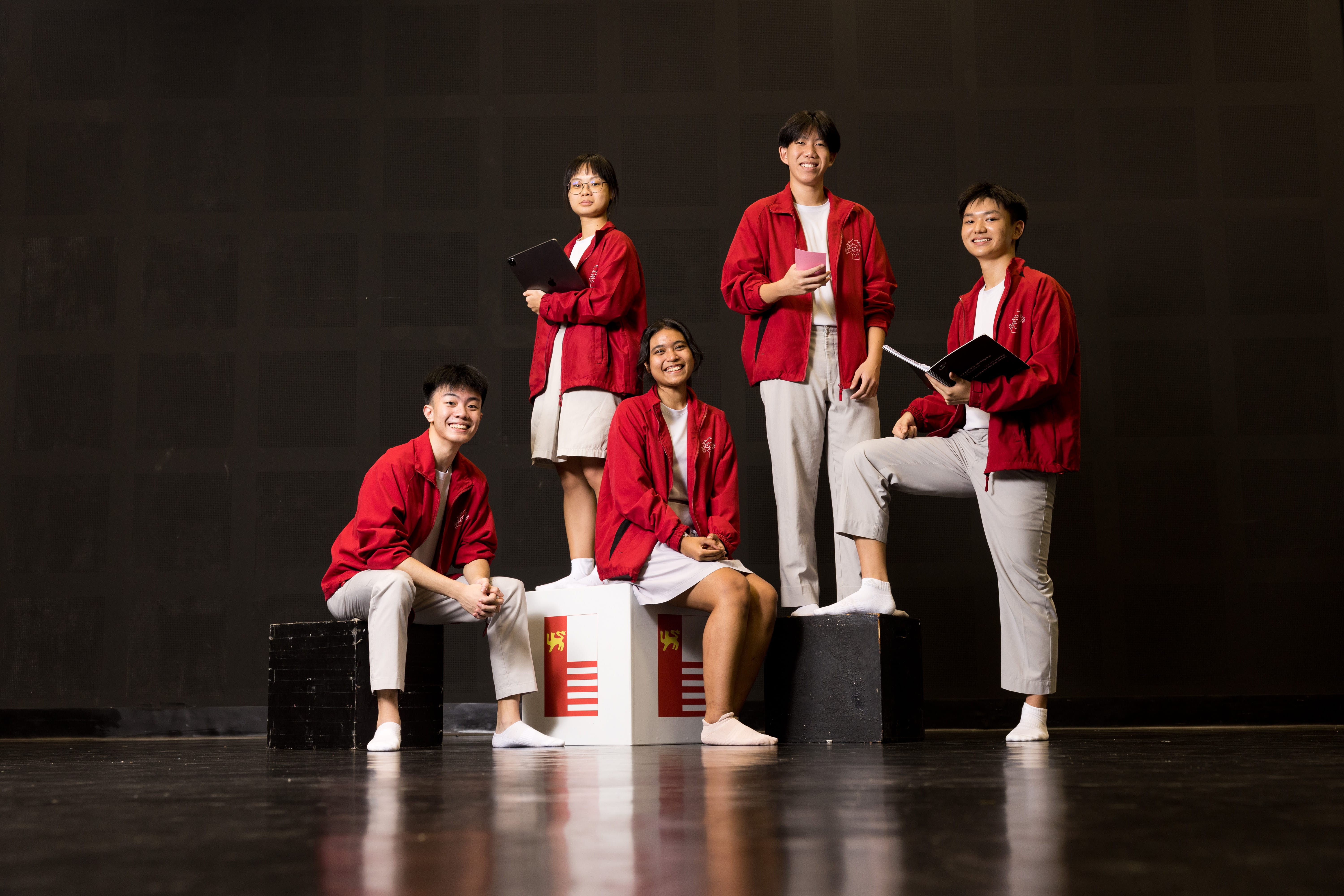 Five students in red jackets and light pants pose on stage, some holding folders and notebooks.