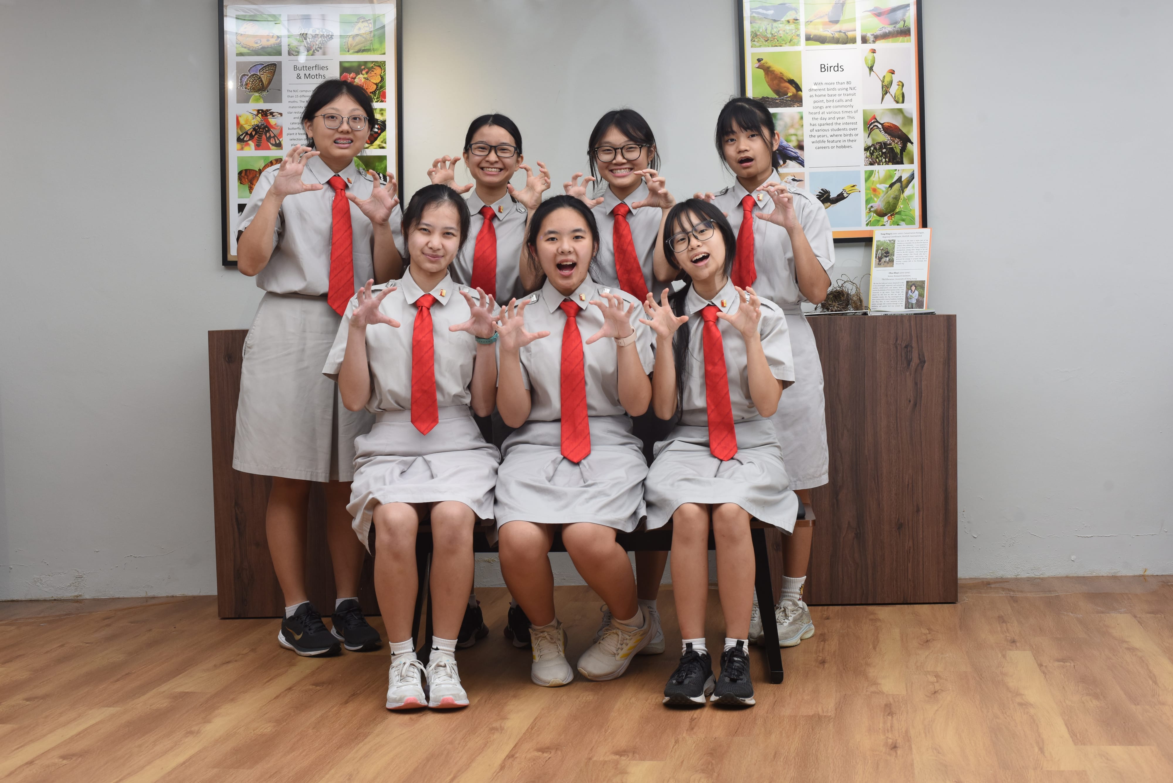 Seven female students in gray uniforms with red ties pose playfully with claw hands in front of nature posters.