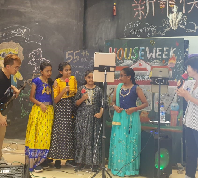Four girls in colorful traditional Indian dresses hold microphones on a stage with a chalkboard reading "House Week."