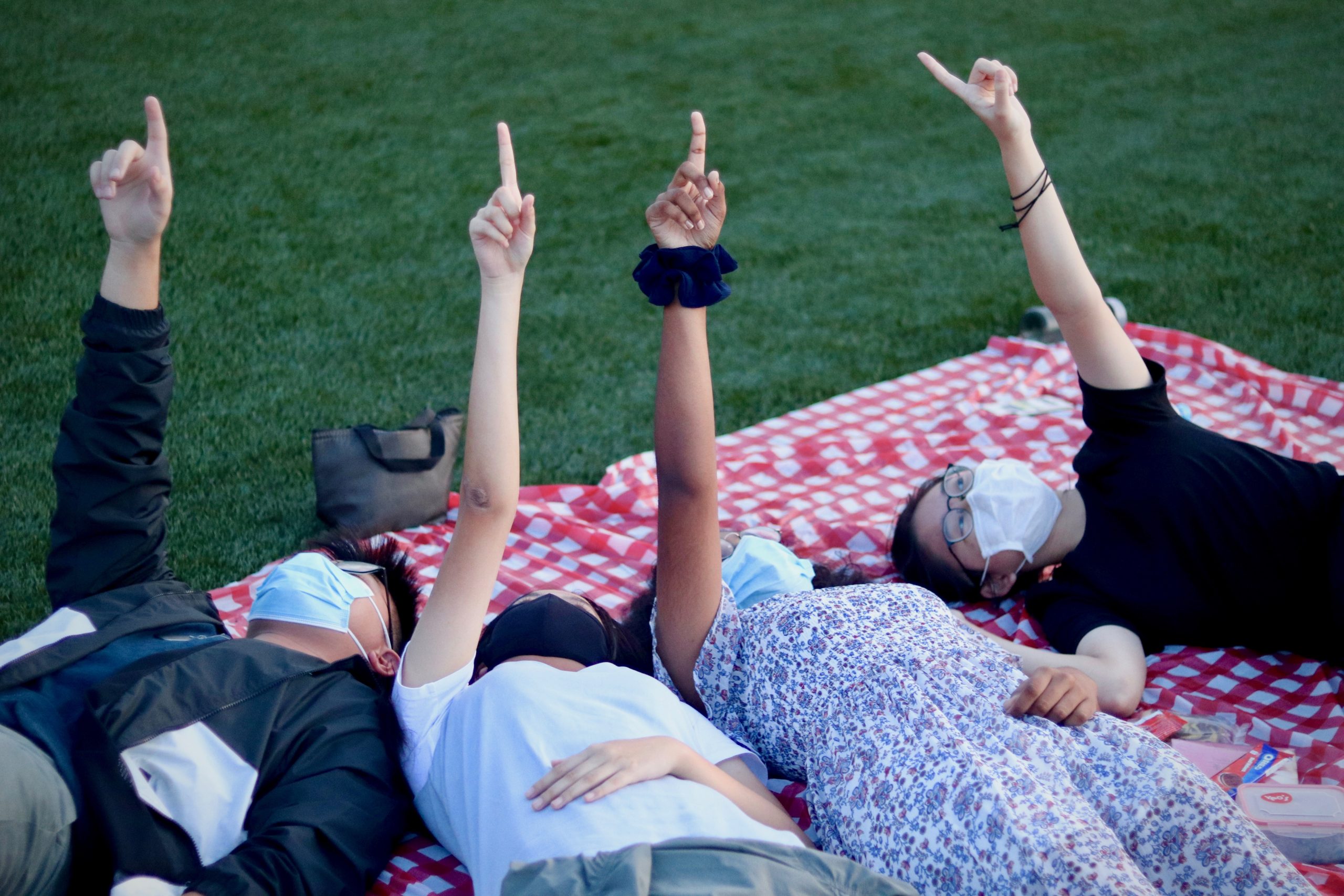 Four people wearing face masks lie on a red checkered blanket on grass, each pointing one finger upward.