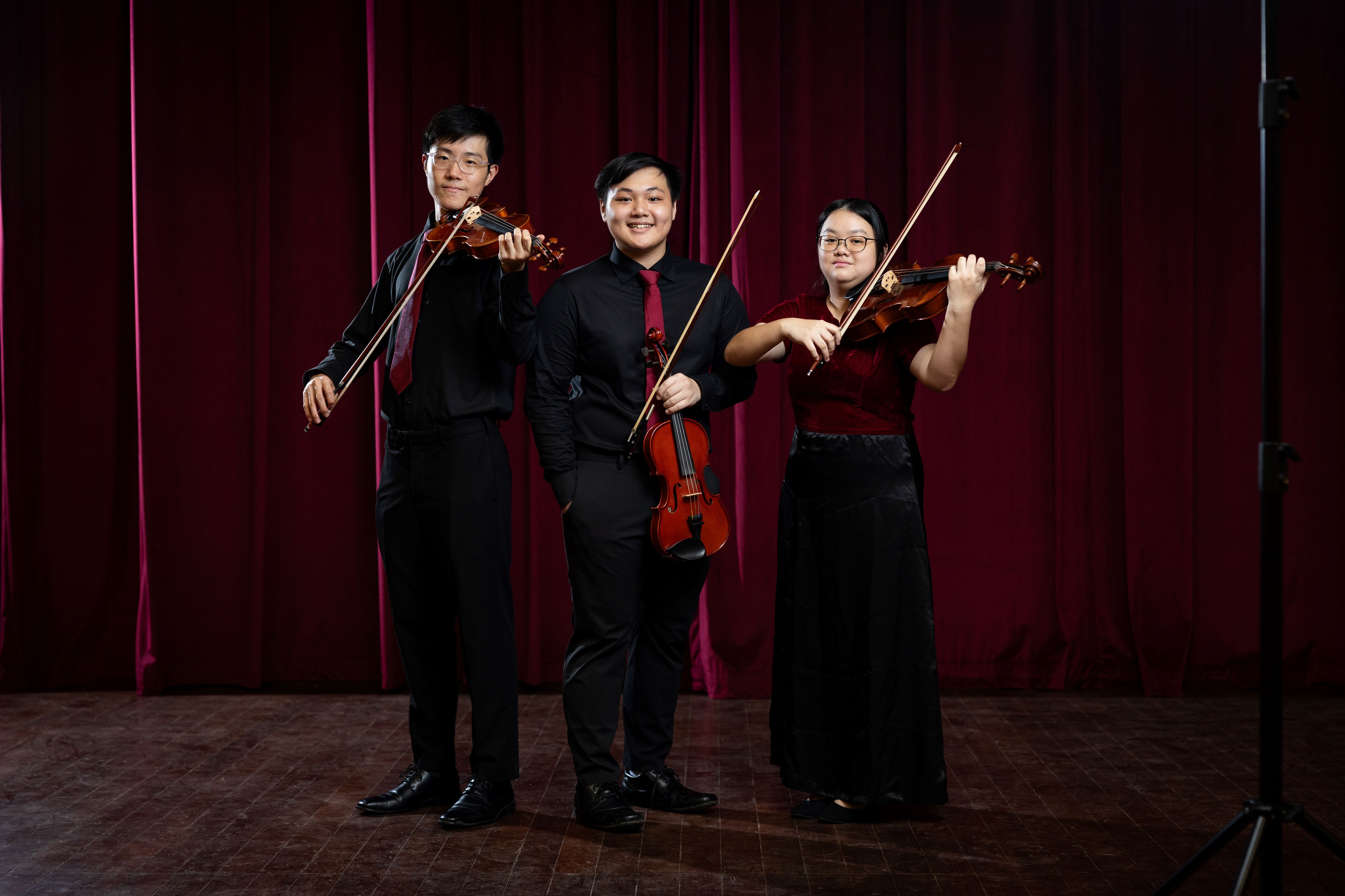 Three young musicians in black formal attire hold violins on a wooden stage in front of a red curtain.