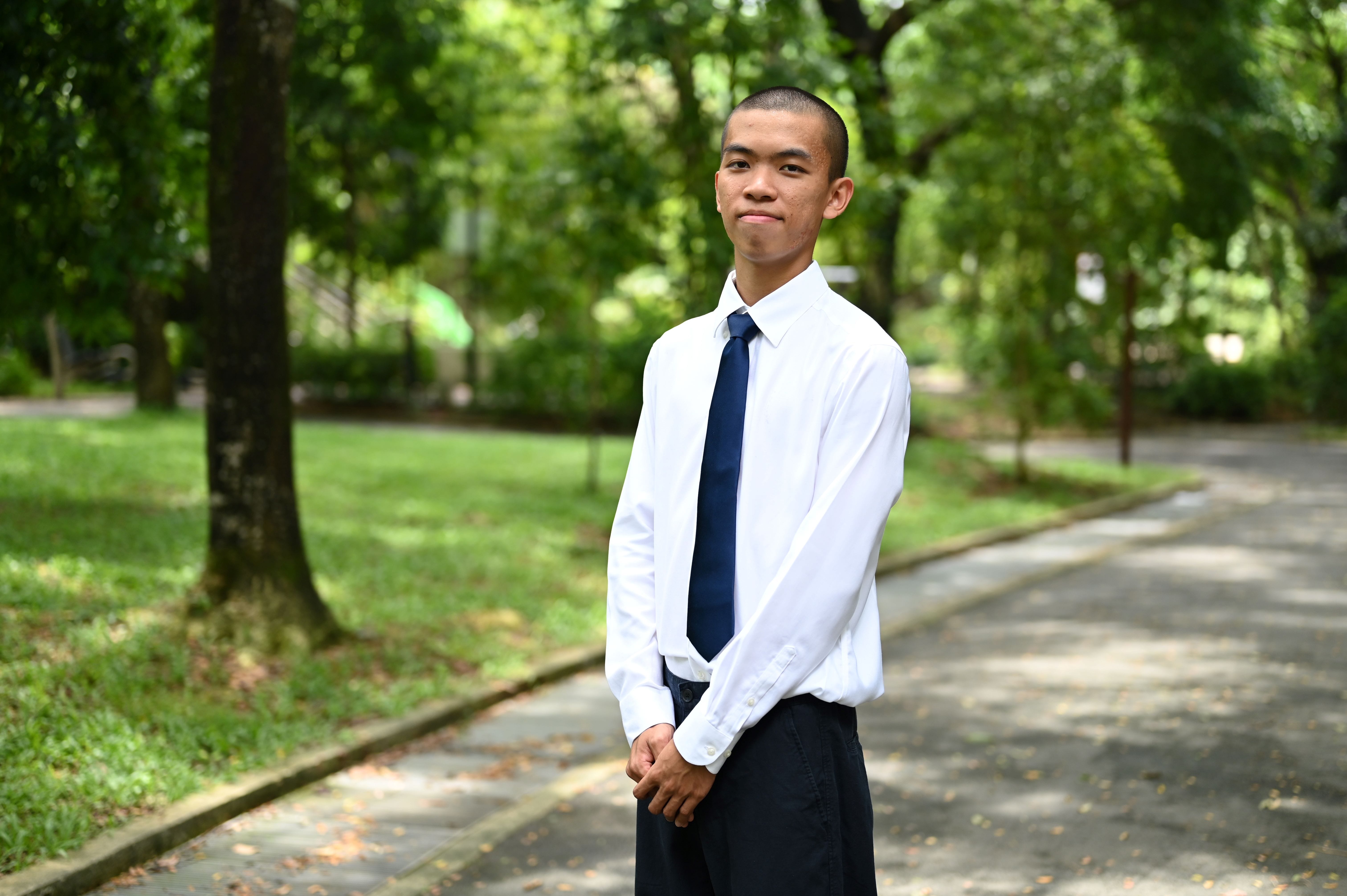 Young man in a white shirt and navy tie standing on a tree-lined park path, smiling slightly.