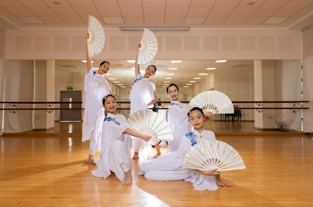 Five young girls in white and blue outfits pose with large white folding fans in a dance studio.